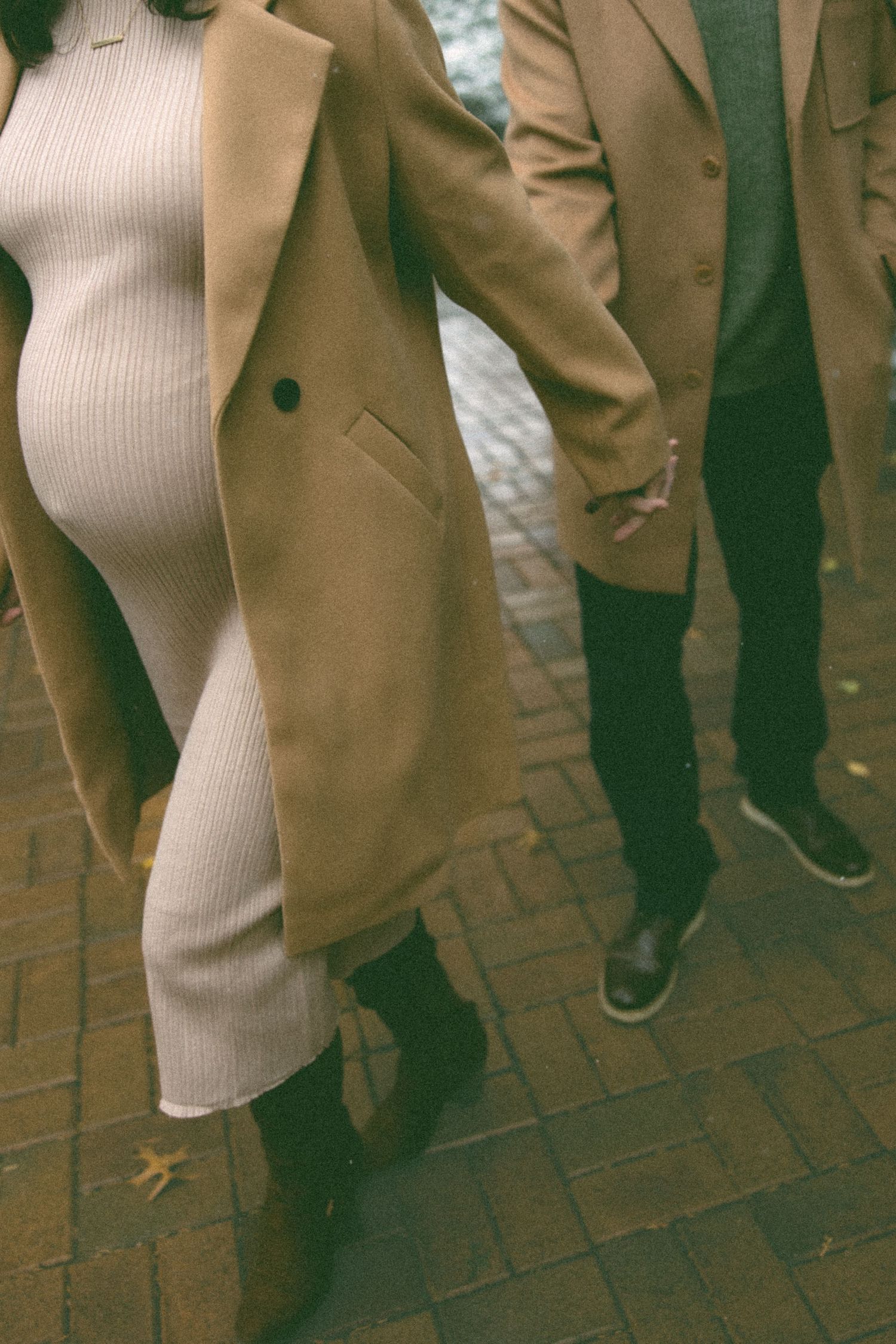 Close-up view of tan coats and white knit dress against brick pavement in winter.