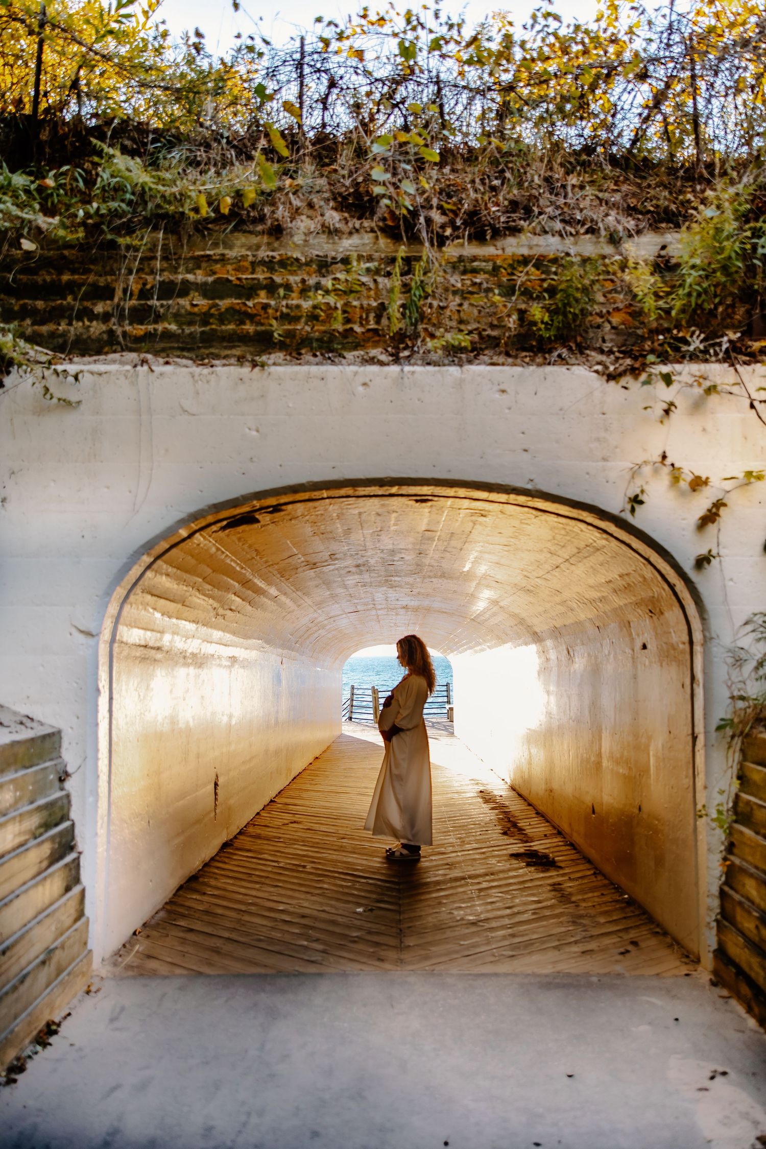 Silhouetted figure stands in bright tunnel with wooden walkway and ivy growing above.
