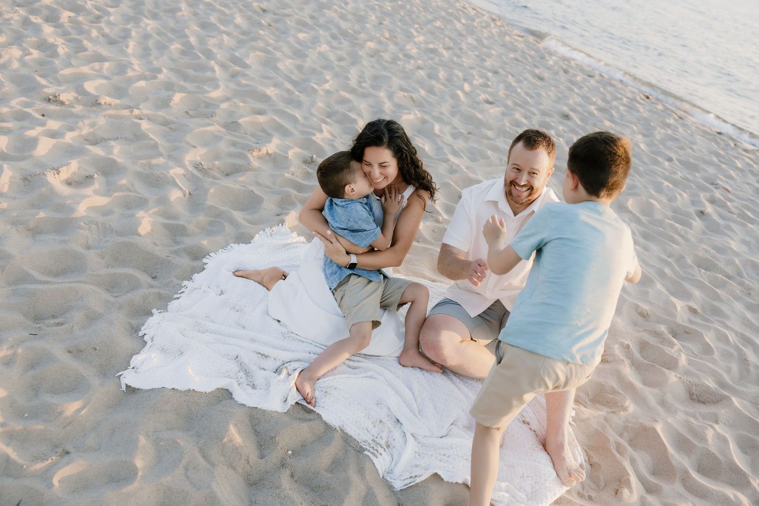 Happy family sitting on white blanket on sandy beach, parents playing with two young children outdoors.
