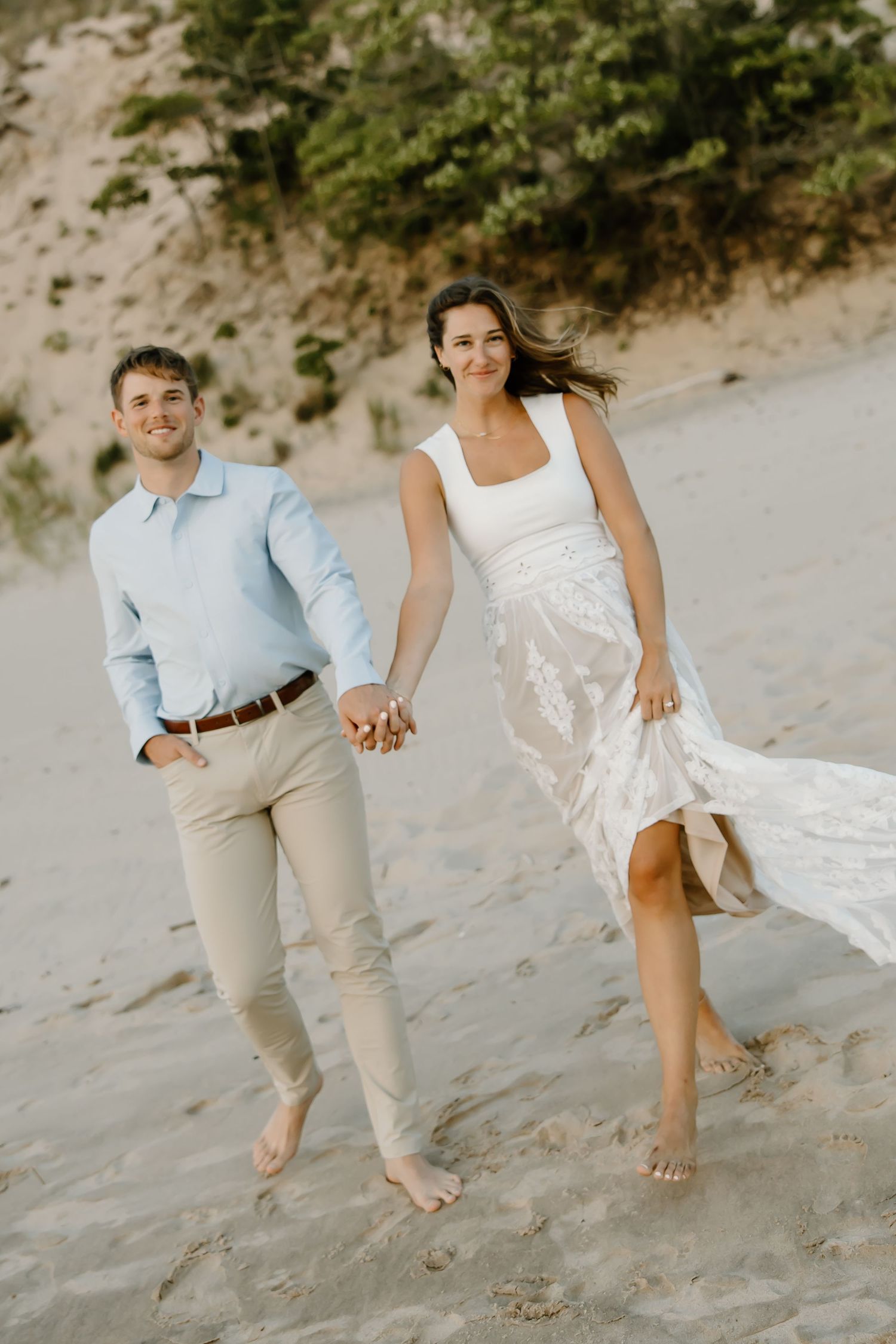 Couple walking barefoot on beach sand, with white dress flowing in the wind.