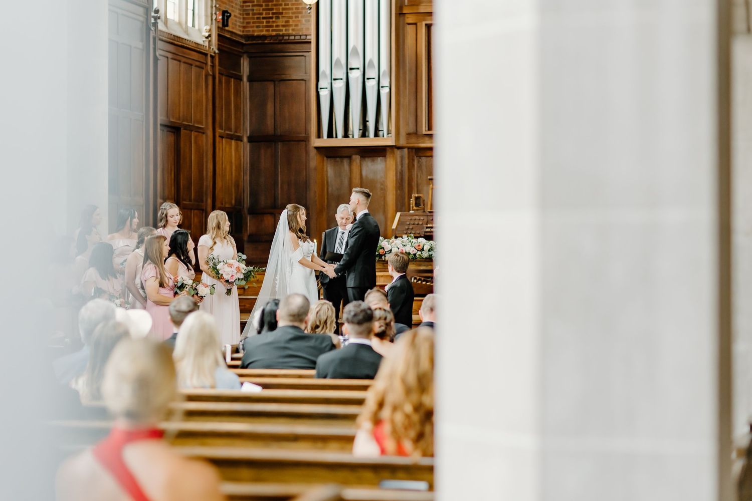 Wedding ceremony in progress inside elegant chapel with white columns and wooden details.