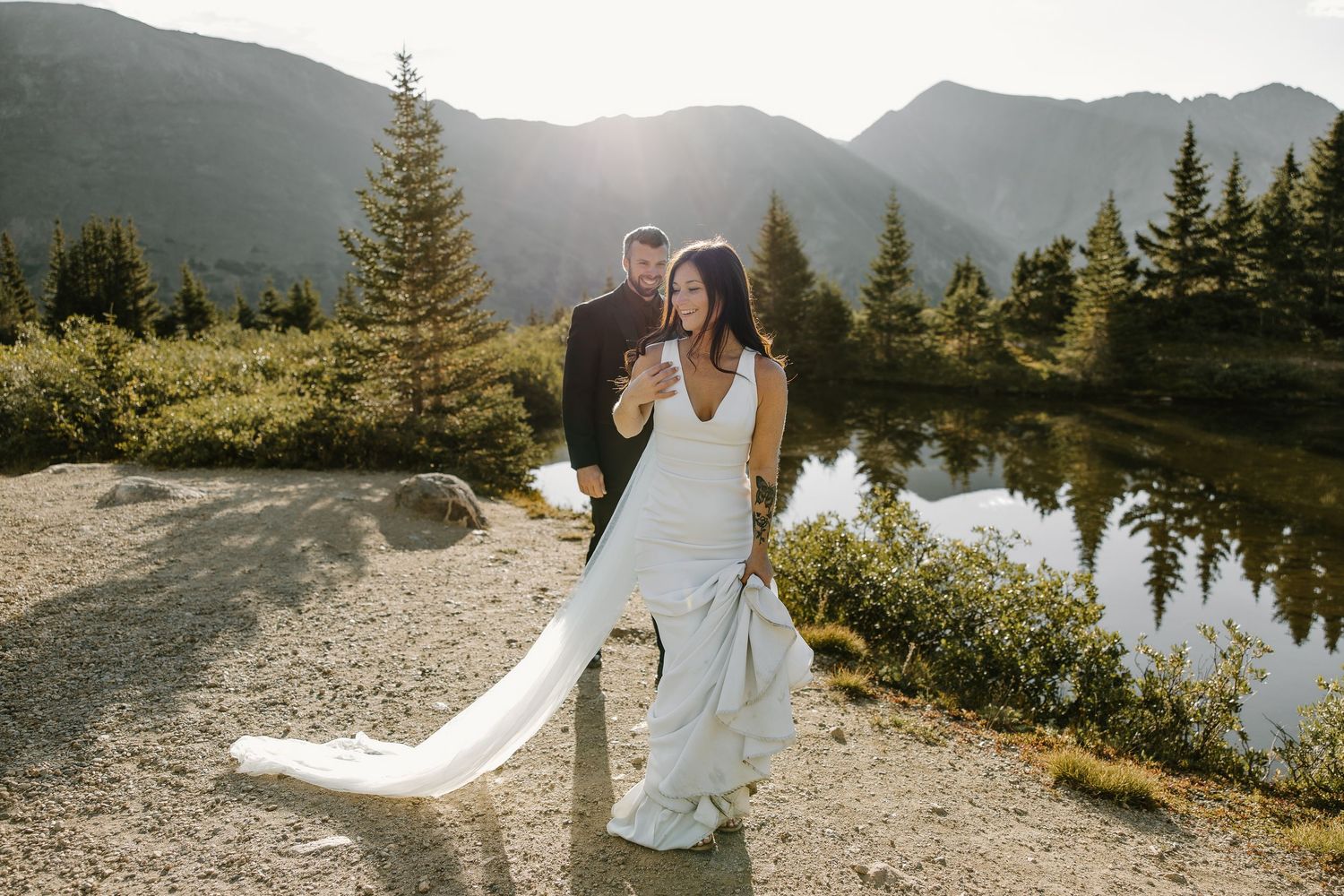 Wedding couple poses near an alpine lake surrounded by mountain peaks and evergreen trees.