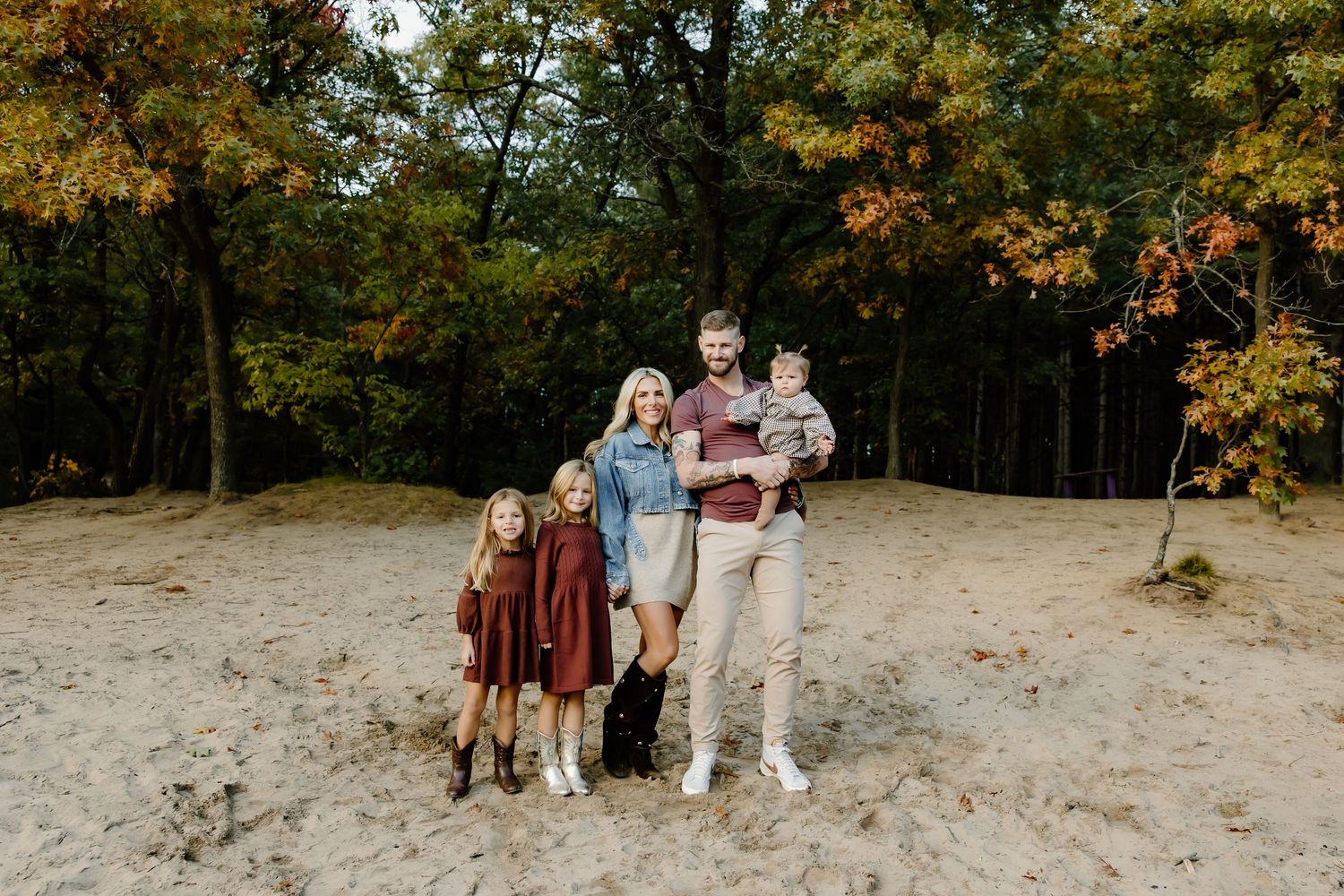 Group portrait of family members standing together on sandy ground with fall foliage in background.