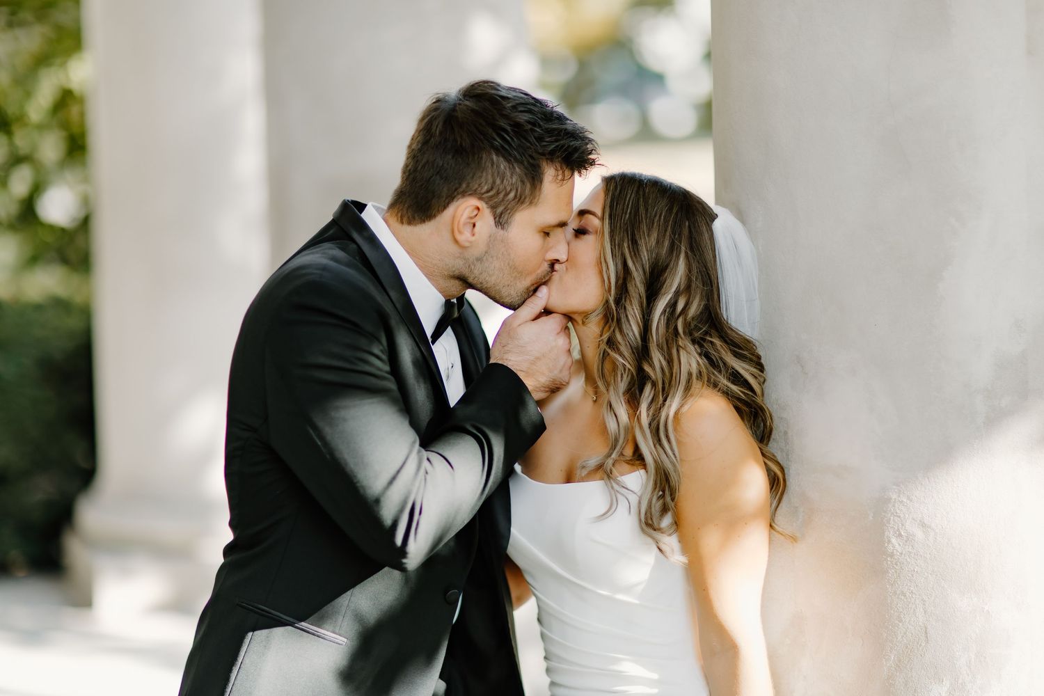 Romantic wedding couple sharing an intimate moment under white columns in natural light.