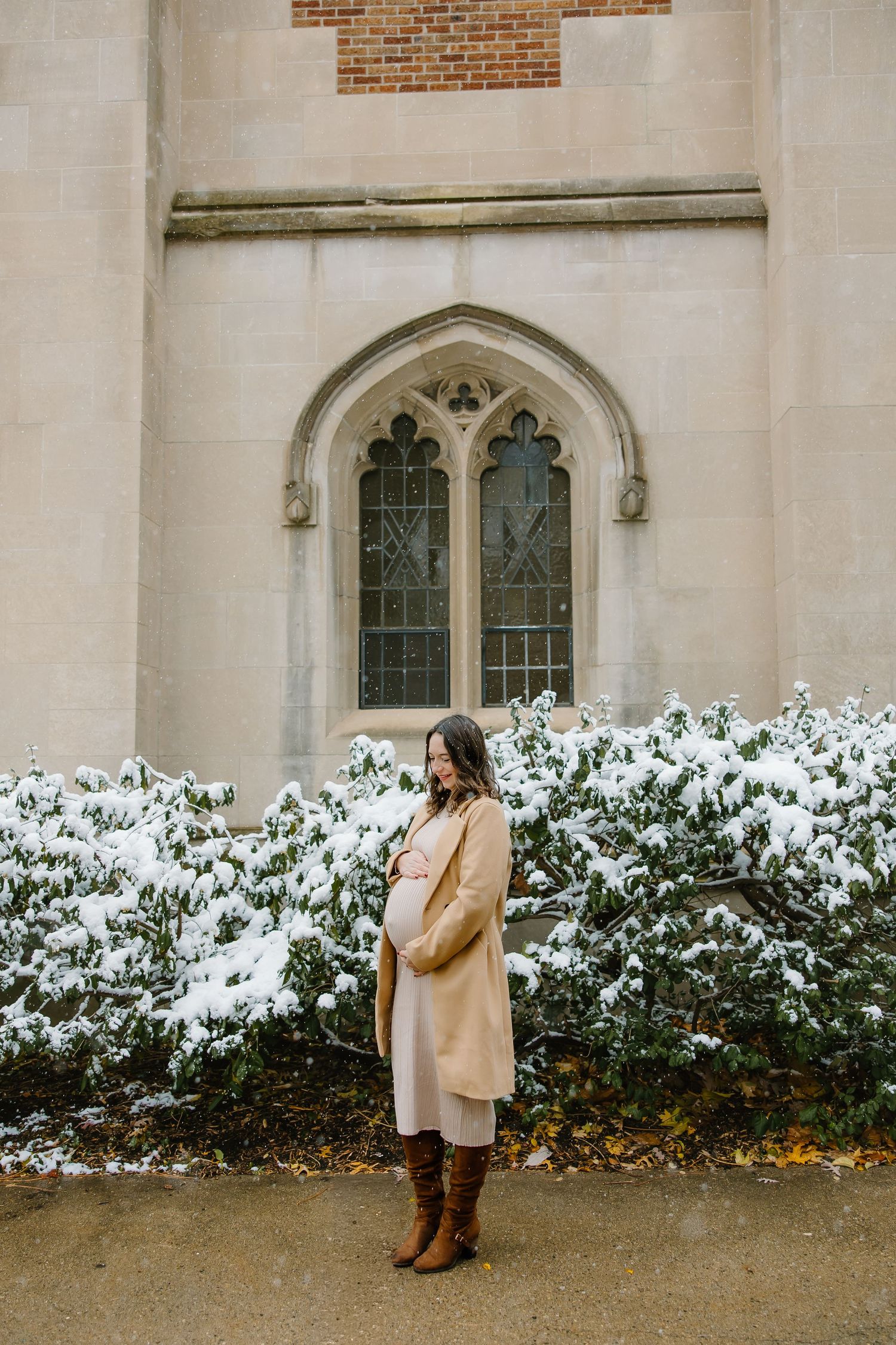 Woman in tan winter coat stands by snowy bushes and Gothic church window.