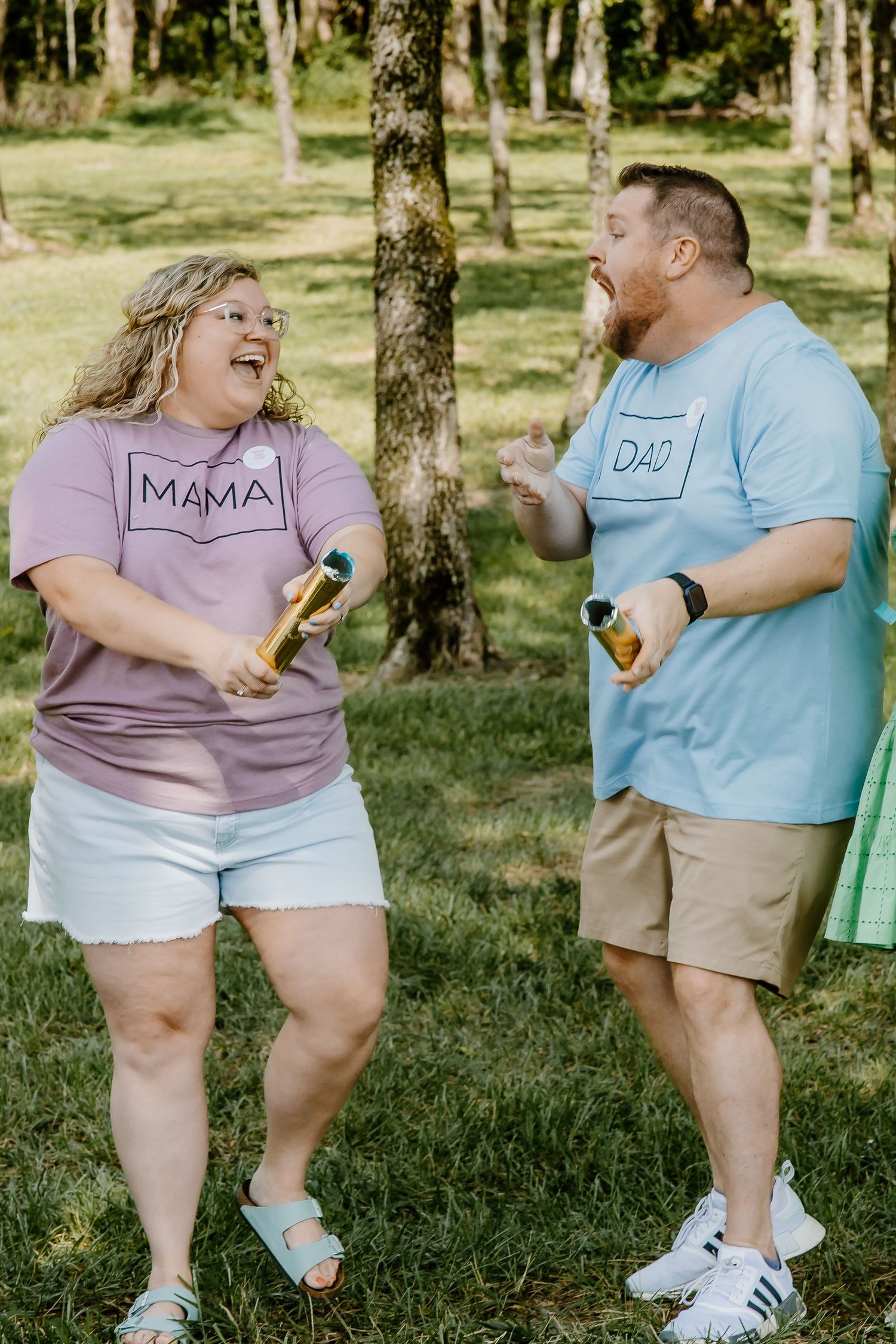 A couple wearing matching pastel shirts playfully pose together in a wooded outdoor setting.