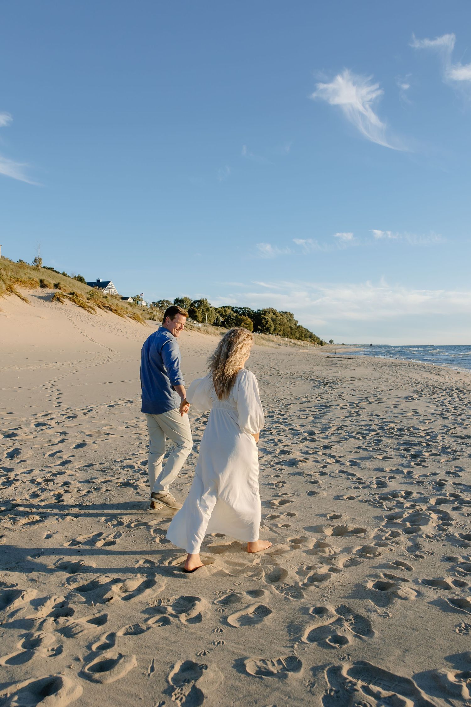 Couple taking a leisurely walk on a sandy beach with dunes in the background.