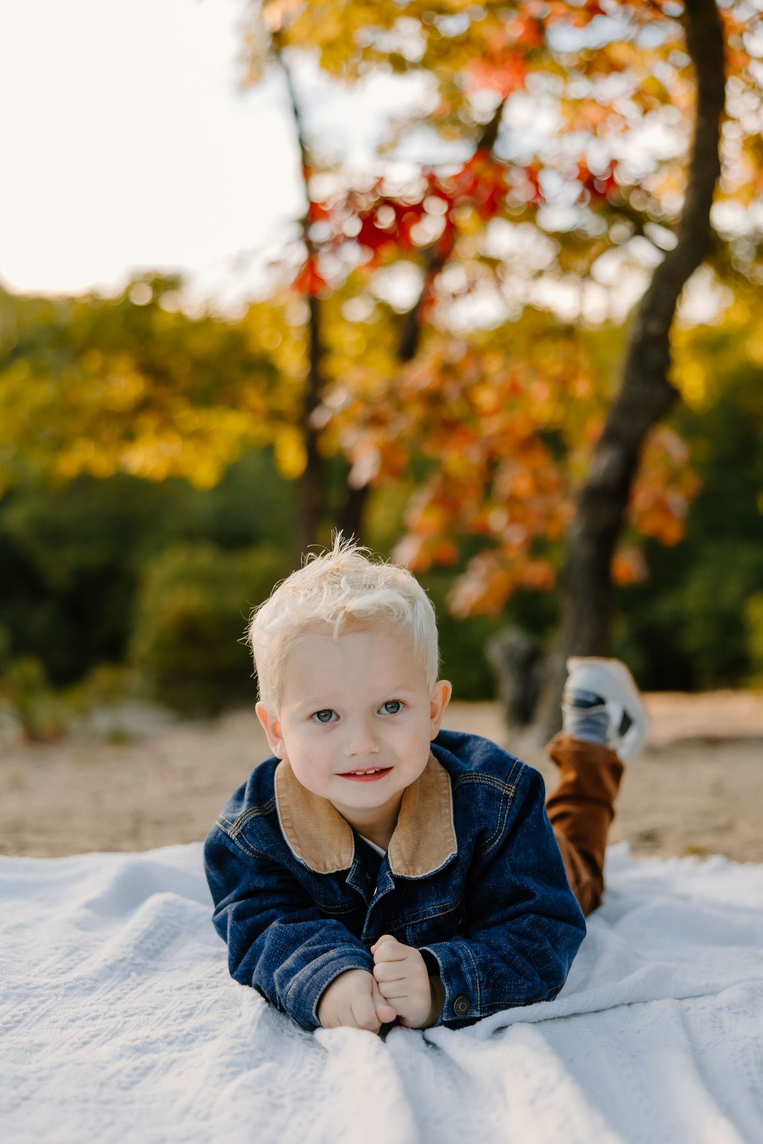 A small child poses on a white blanket with autumn foliage in the background.