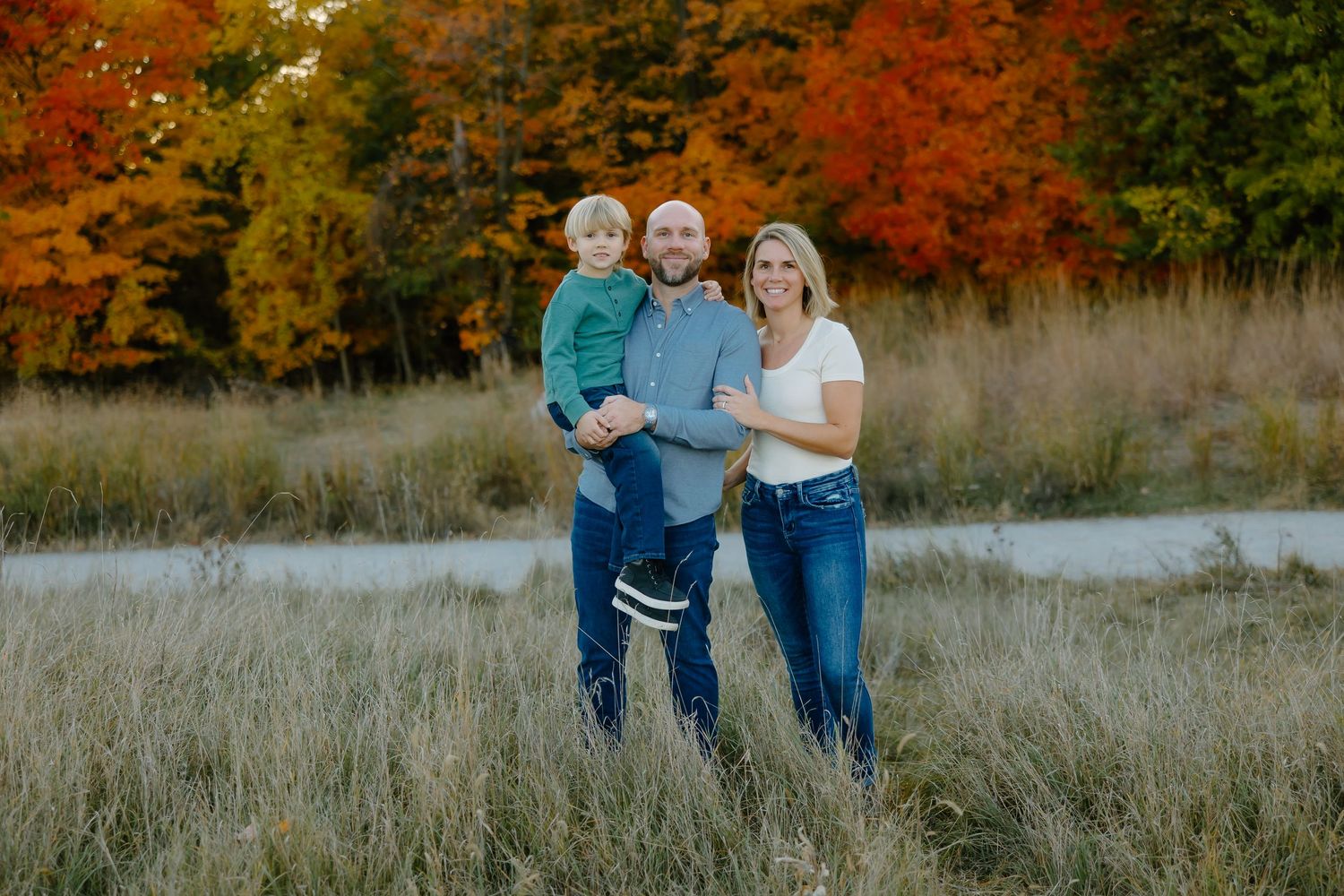 Family enjoying a fall day together in a field with beautiful orange foliage in the background.
