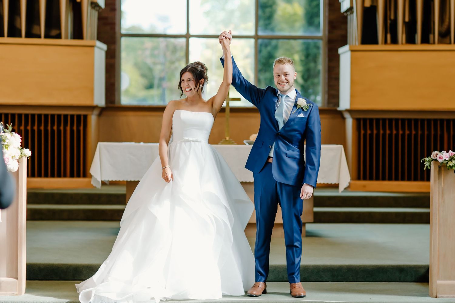 Newlyweds celebrate with raised hands at the altar of a sunlit church sanctuary.