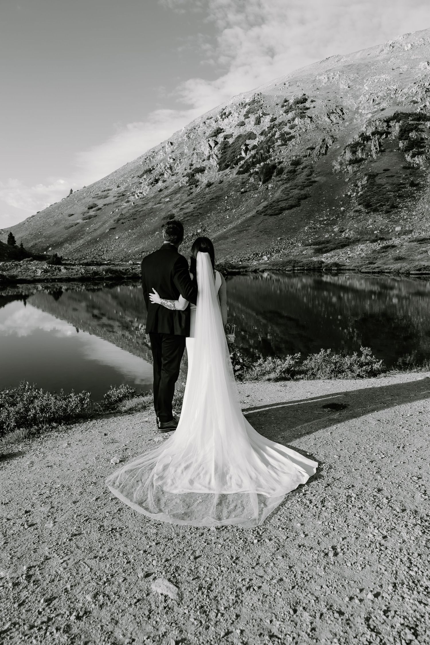 Black and white photo of newlyweds by a serene mountain lake with dramatic reflections.