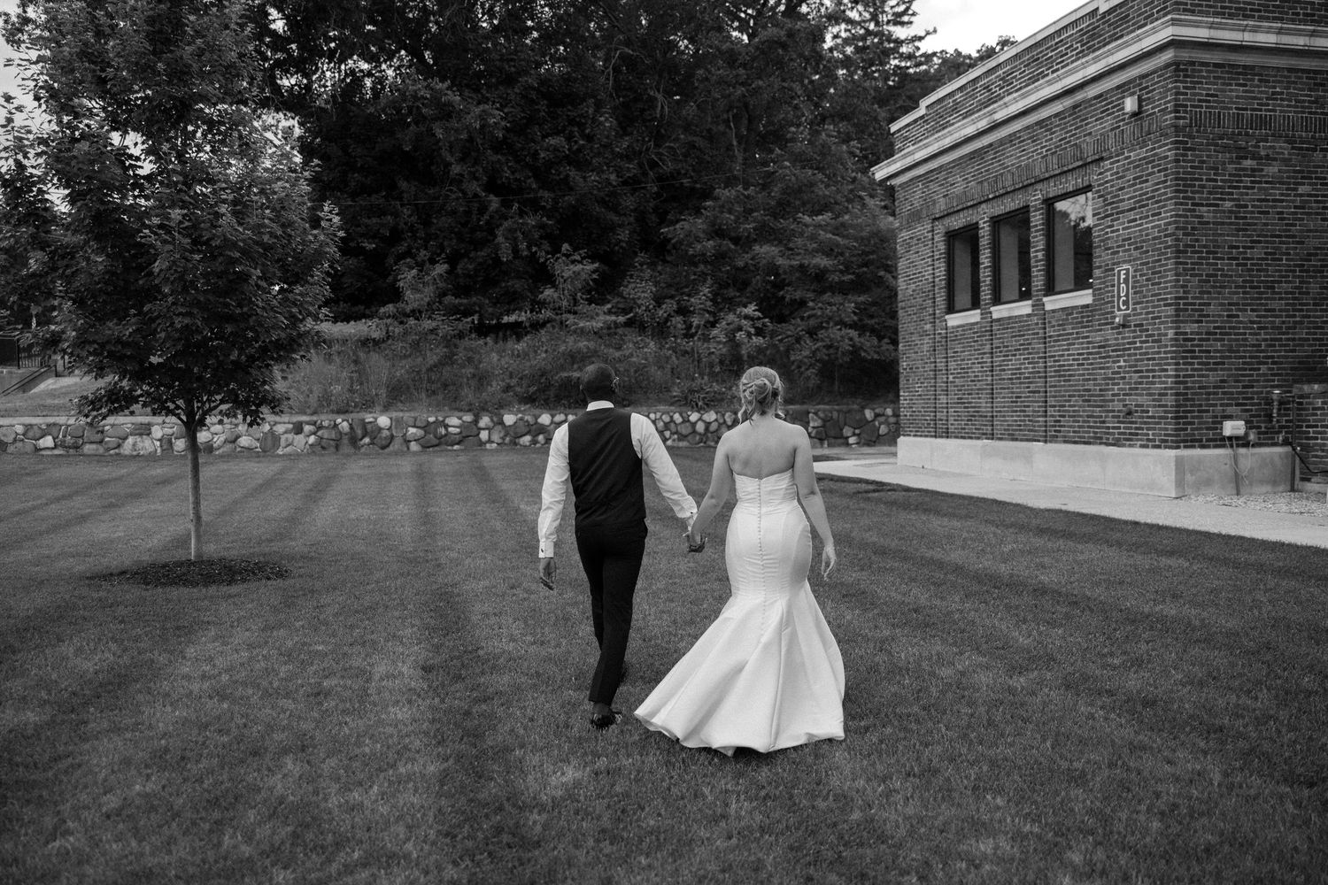 A romantic wedding couple walks across a grassy hill near a brick building on their special day.