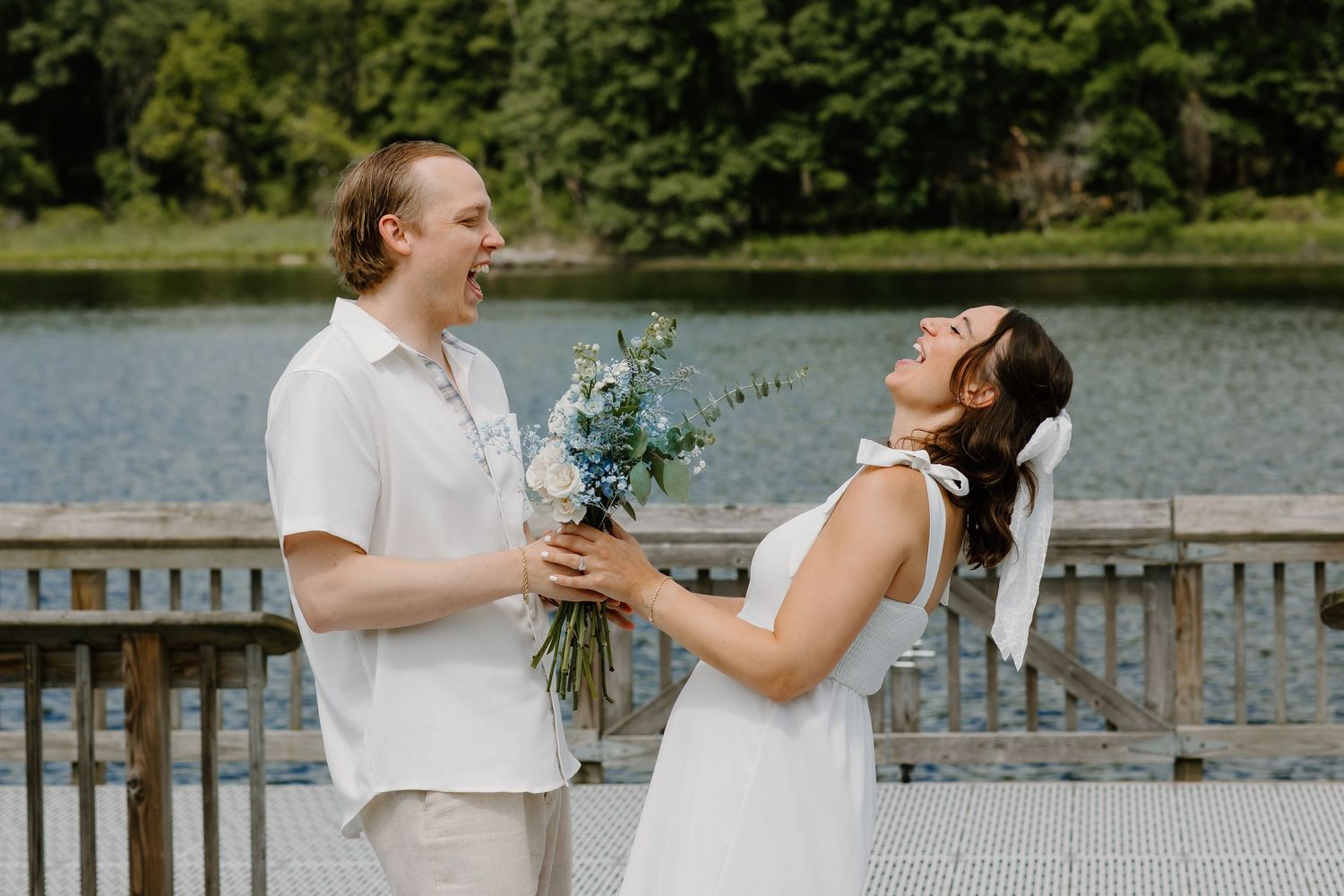 Couple sharing intimate moment on wooden dock by lake with blue flower bouquet.