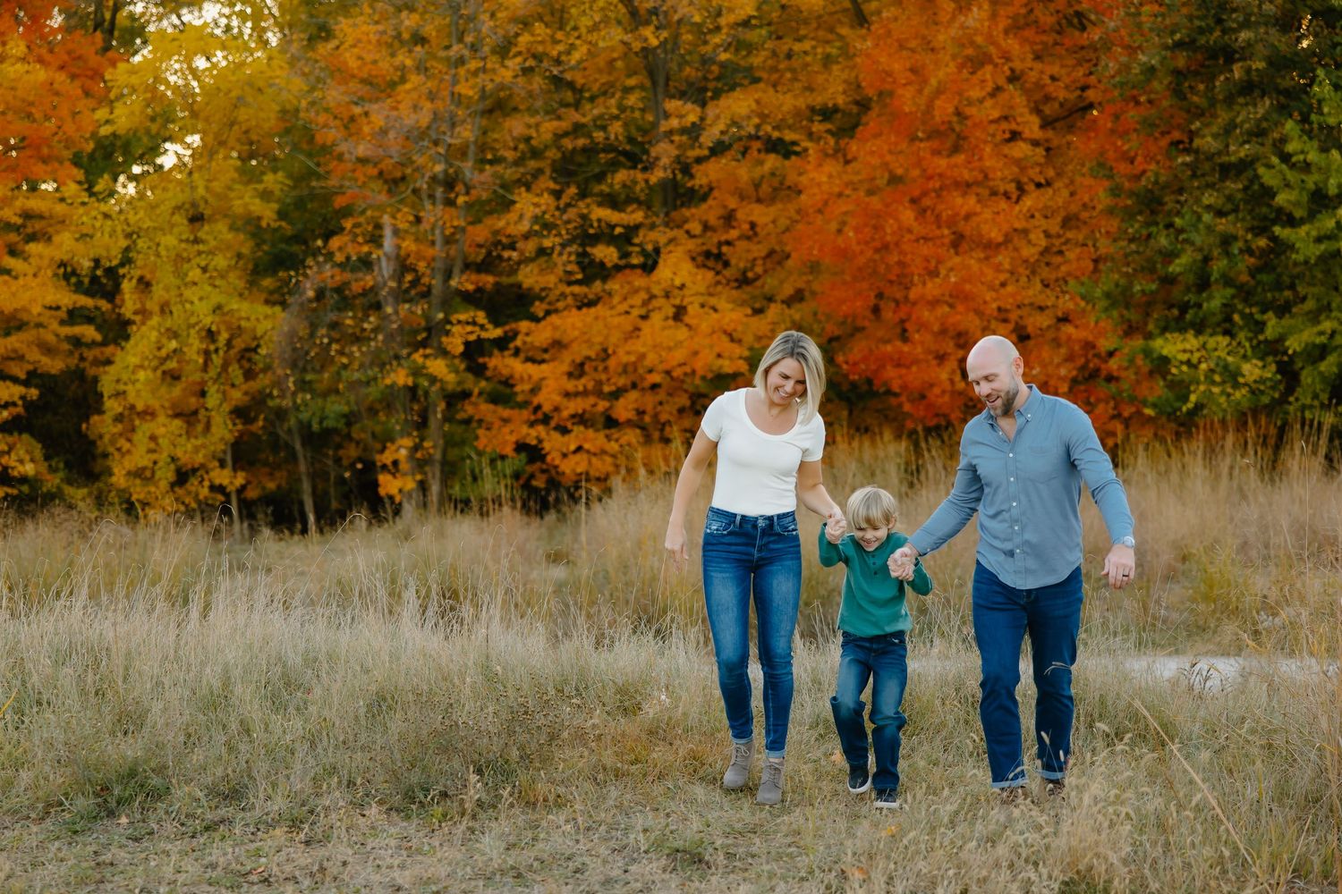 Family of three walks together through an autumn meadow with vibrant orange fall foliage in the background.