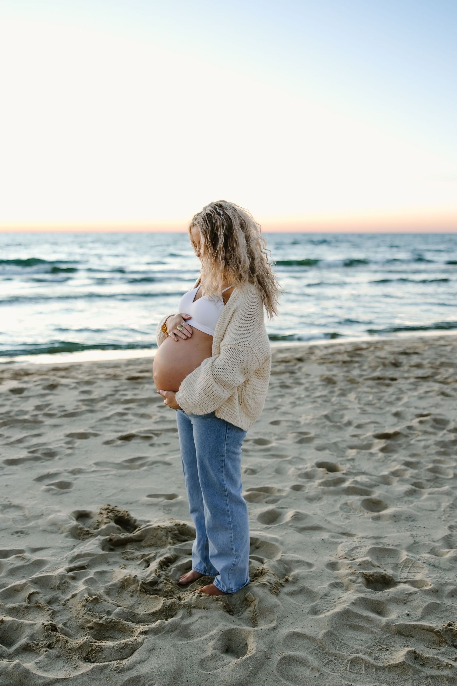 Child in cream sweater and jeans plays on sandy beach at sunset with ocean waves in background.