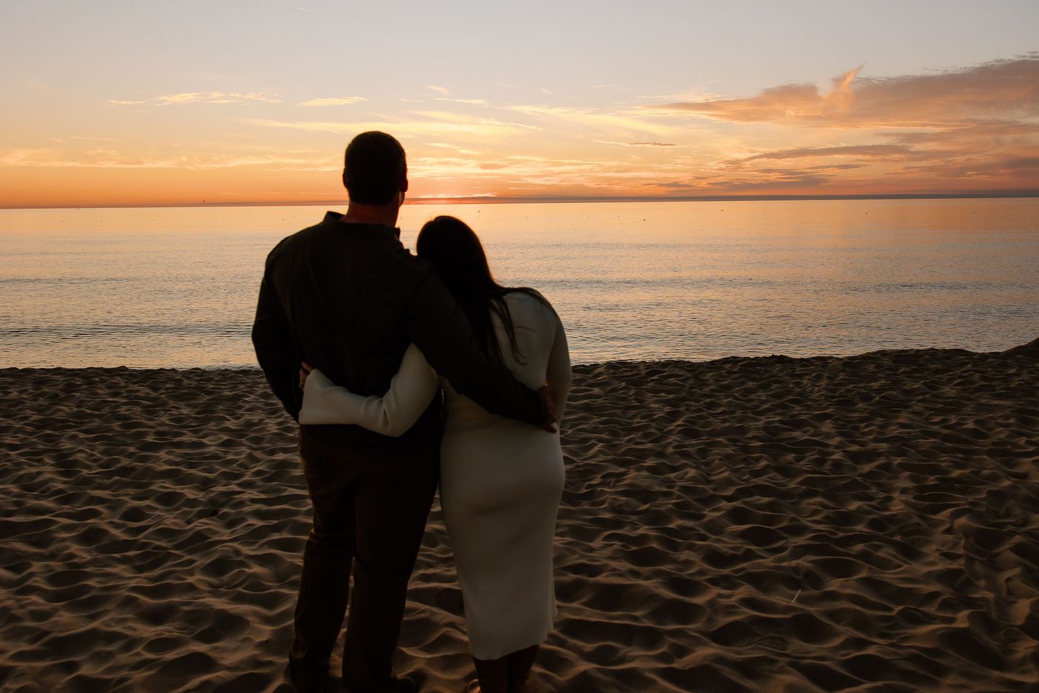Silhouetted couple embracing on beach at sunset with orange sky and ocean backdrop.