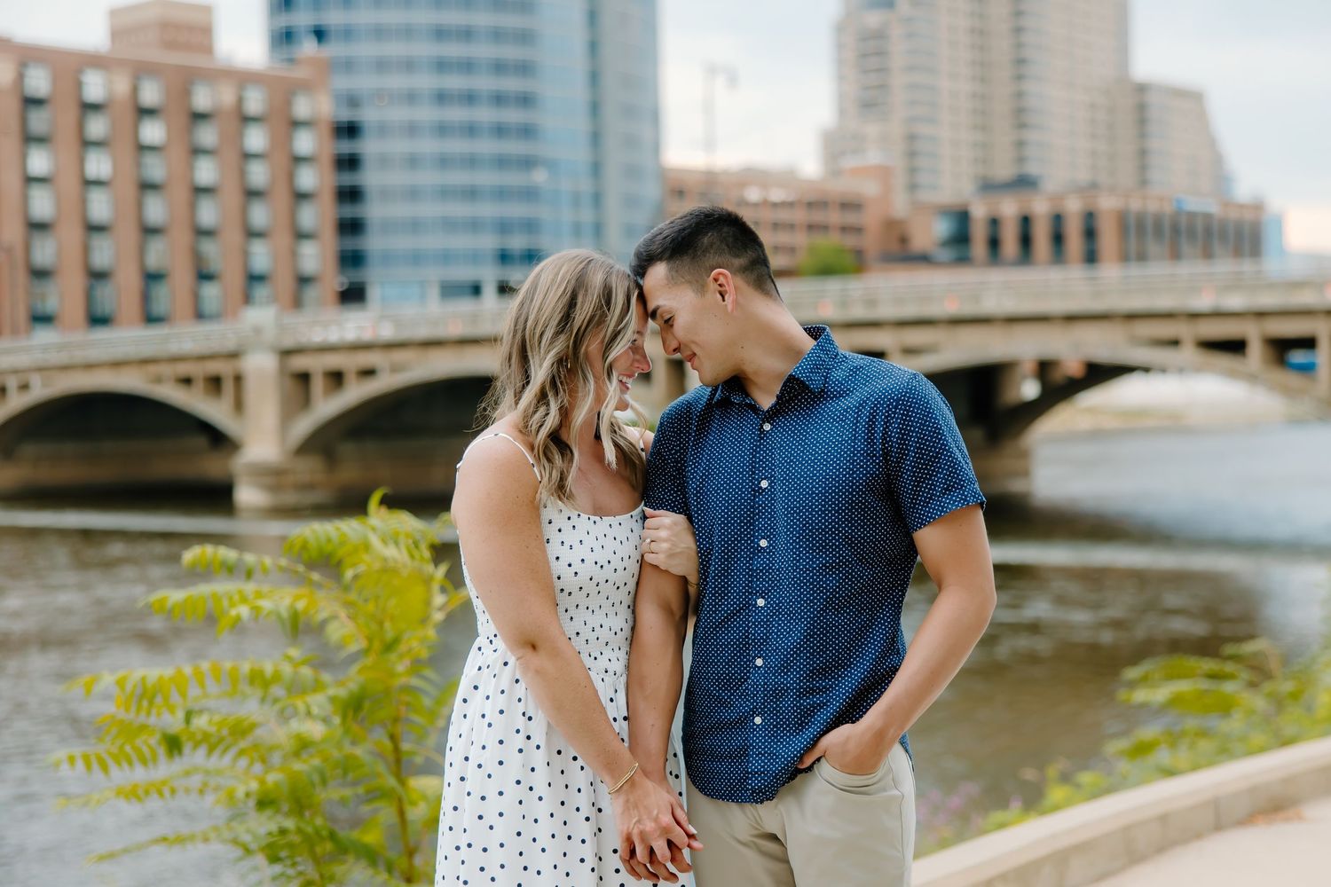 Couple walking along urban riverfront with modern buildings and bridge in background.