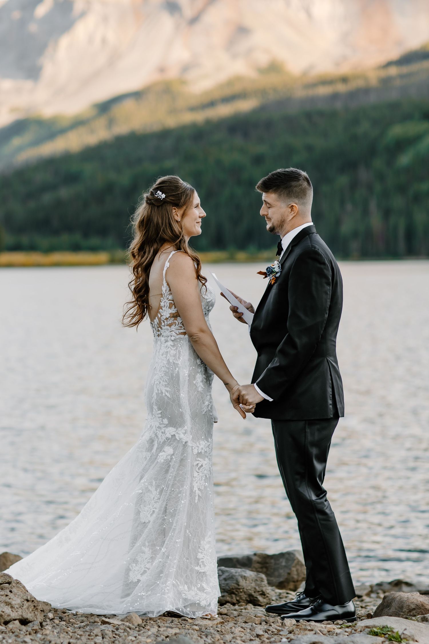 A lakeside wedding ceremony with mountain views as couple exchanges vows on rocky shoreline.