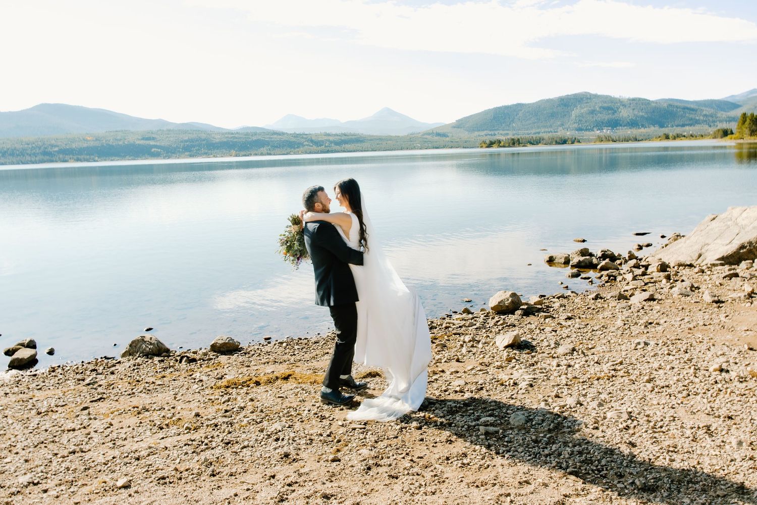 Panoramic view of lake shore with mountains and couple in wedding attire.