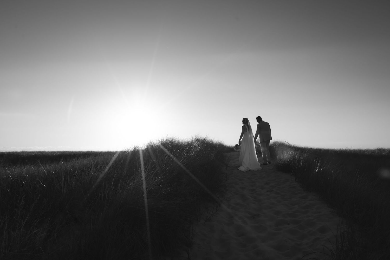 Black and white photo of a couple holding hands on a sandy path at sunset with grass fields in the background.