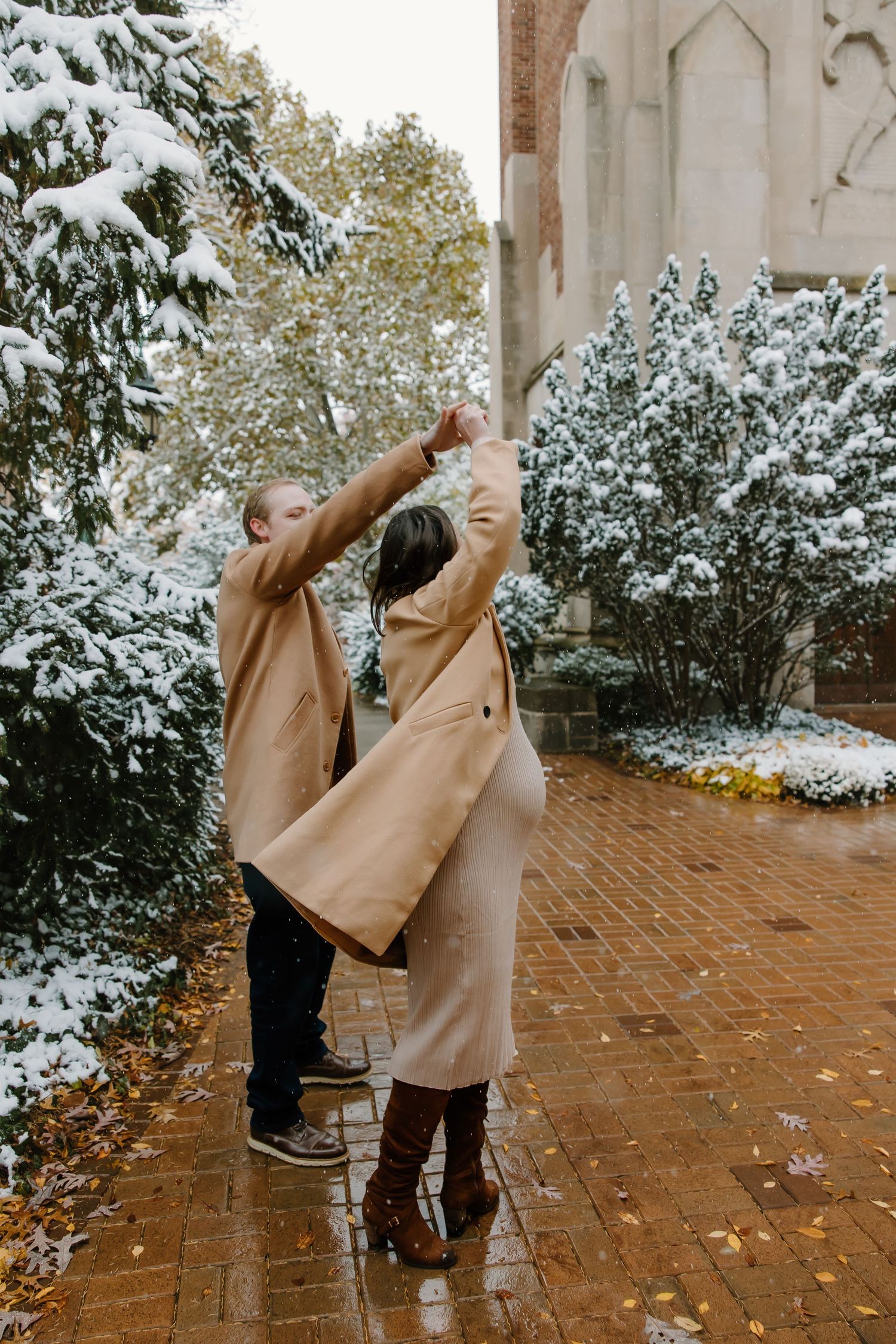 Couple in matching camel coats dancing joyfully on brick pathway surrounded by snow-covered trees.