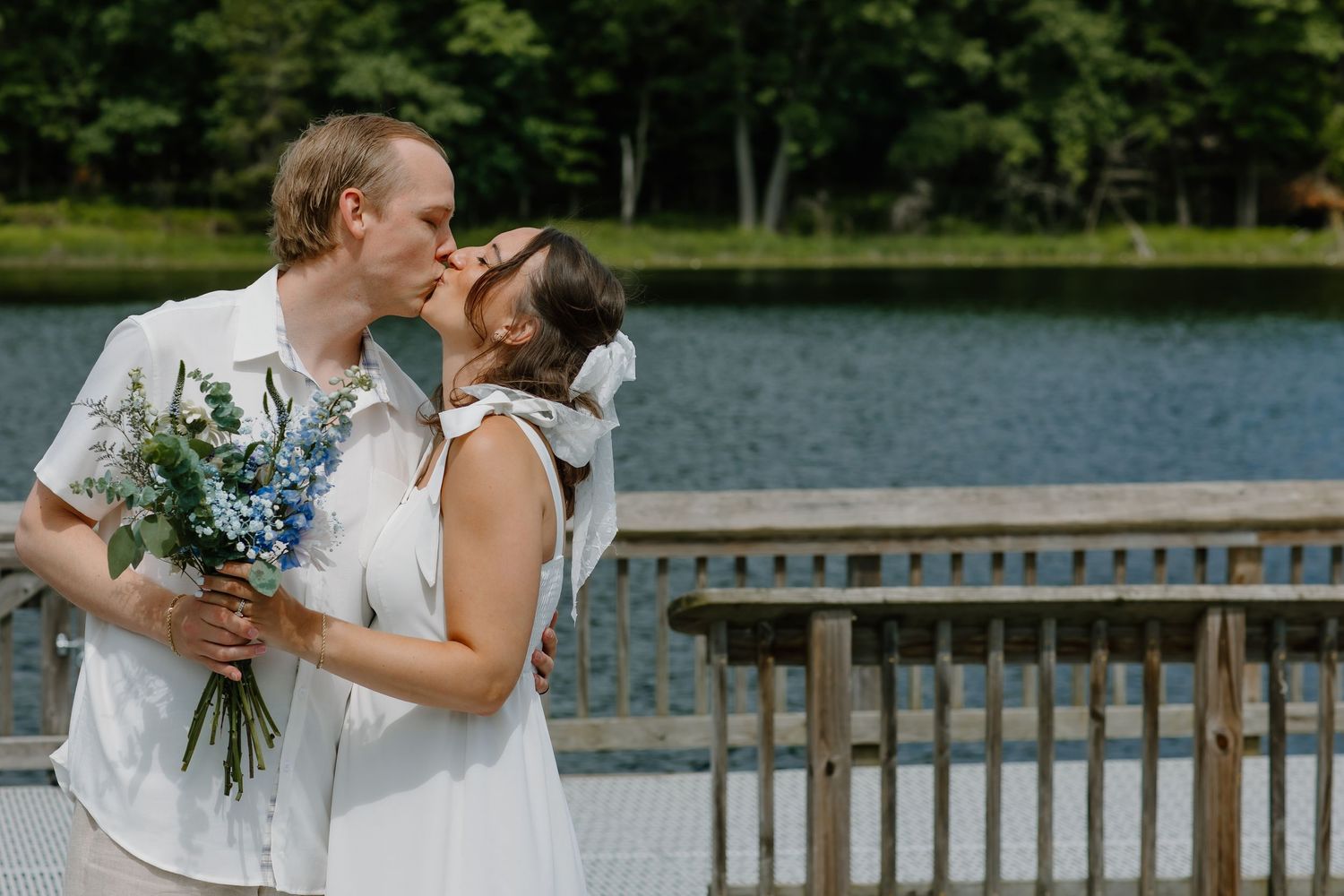 Couple in white outfits sharing a romantic moment by a lake with trees in the background.