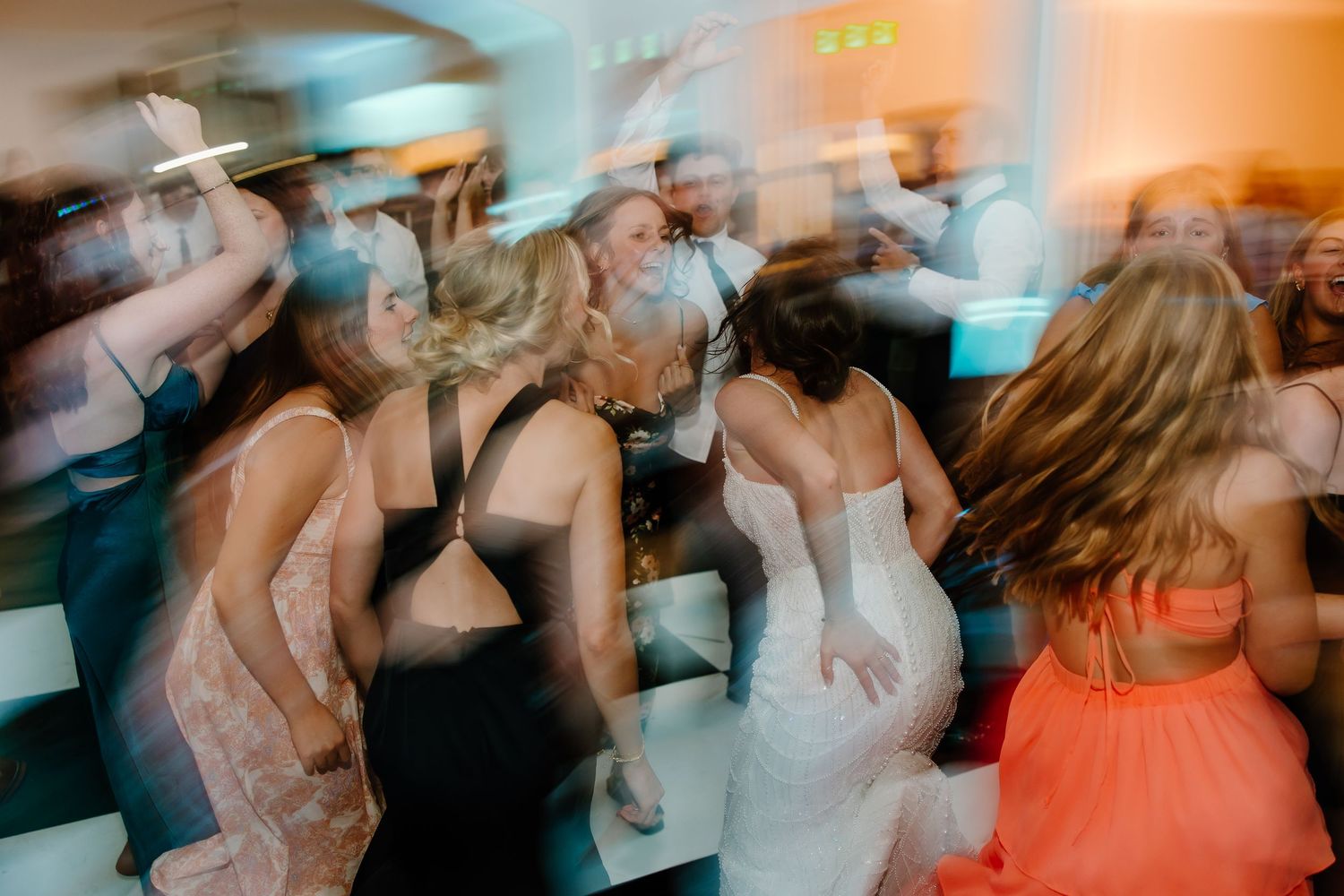 Blurred motion shot of wedding guests dancing energetically at reception with colorful lighting.