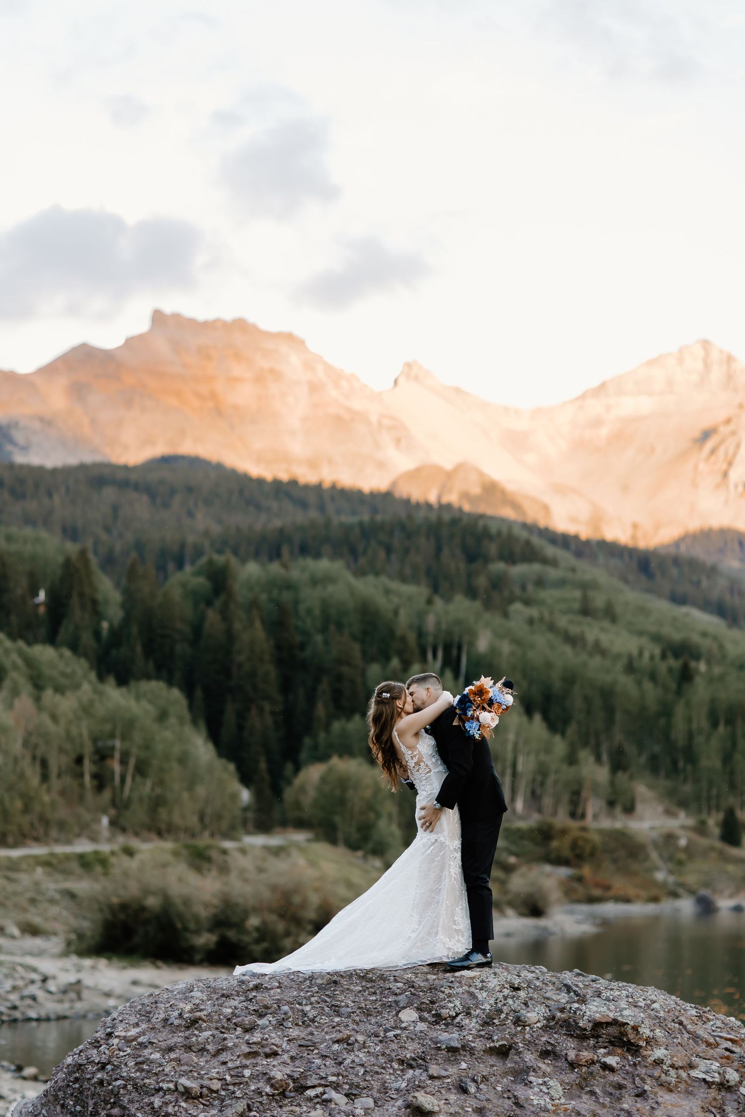 Romantic wedding portrait of a couple kissing on a rock overlooking a mountain lake at sunset.