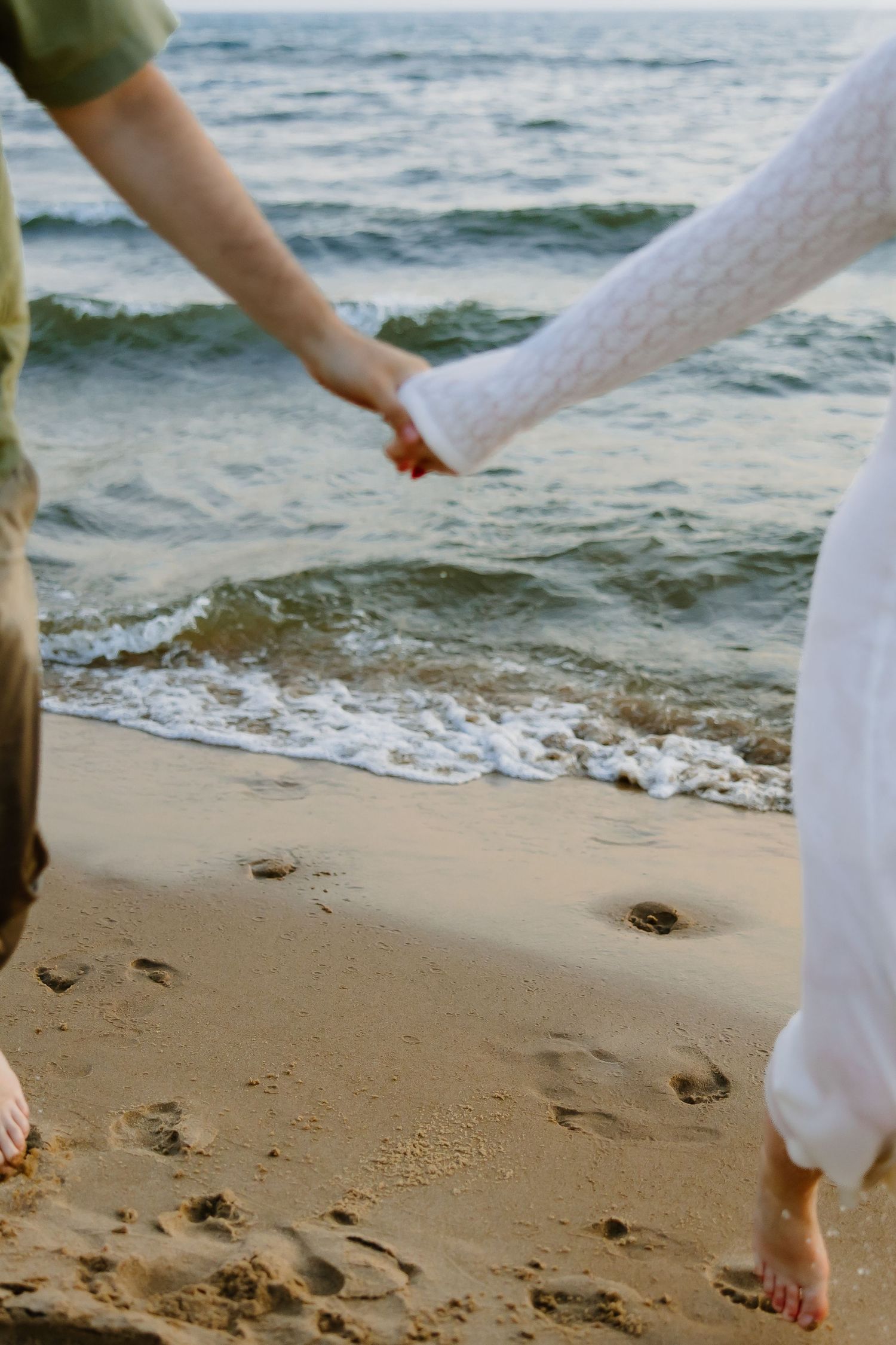Close-up of couple holding hands while walking barefoot on sandy beach with ocean waves.