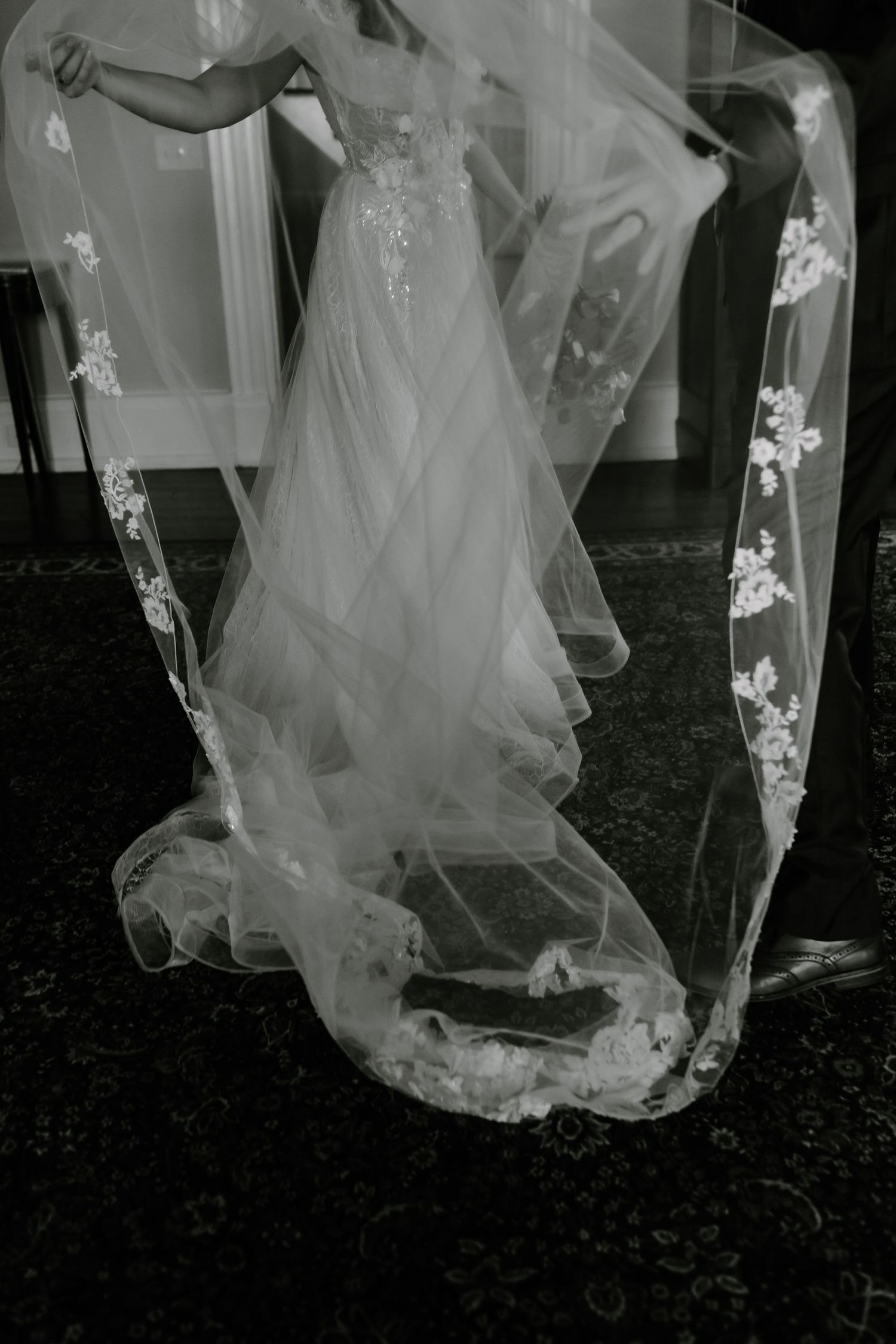 Close-up details of a flowing white wedding dress train on dark patterned carpet.