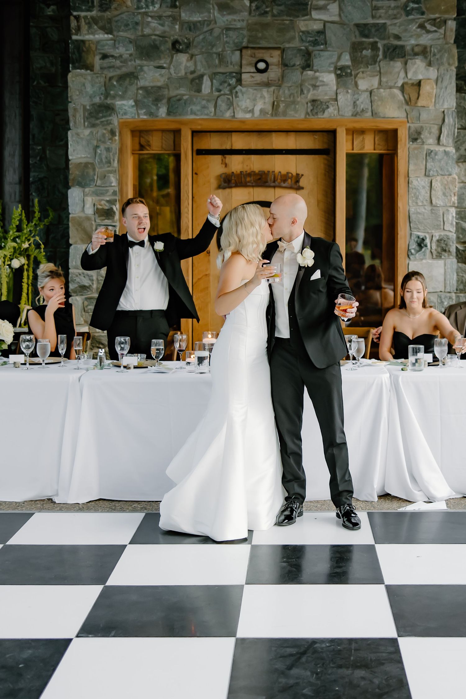 Wedding couple dances on black and white checkered floor against stone wall backdrop.
