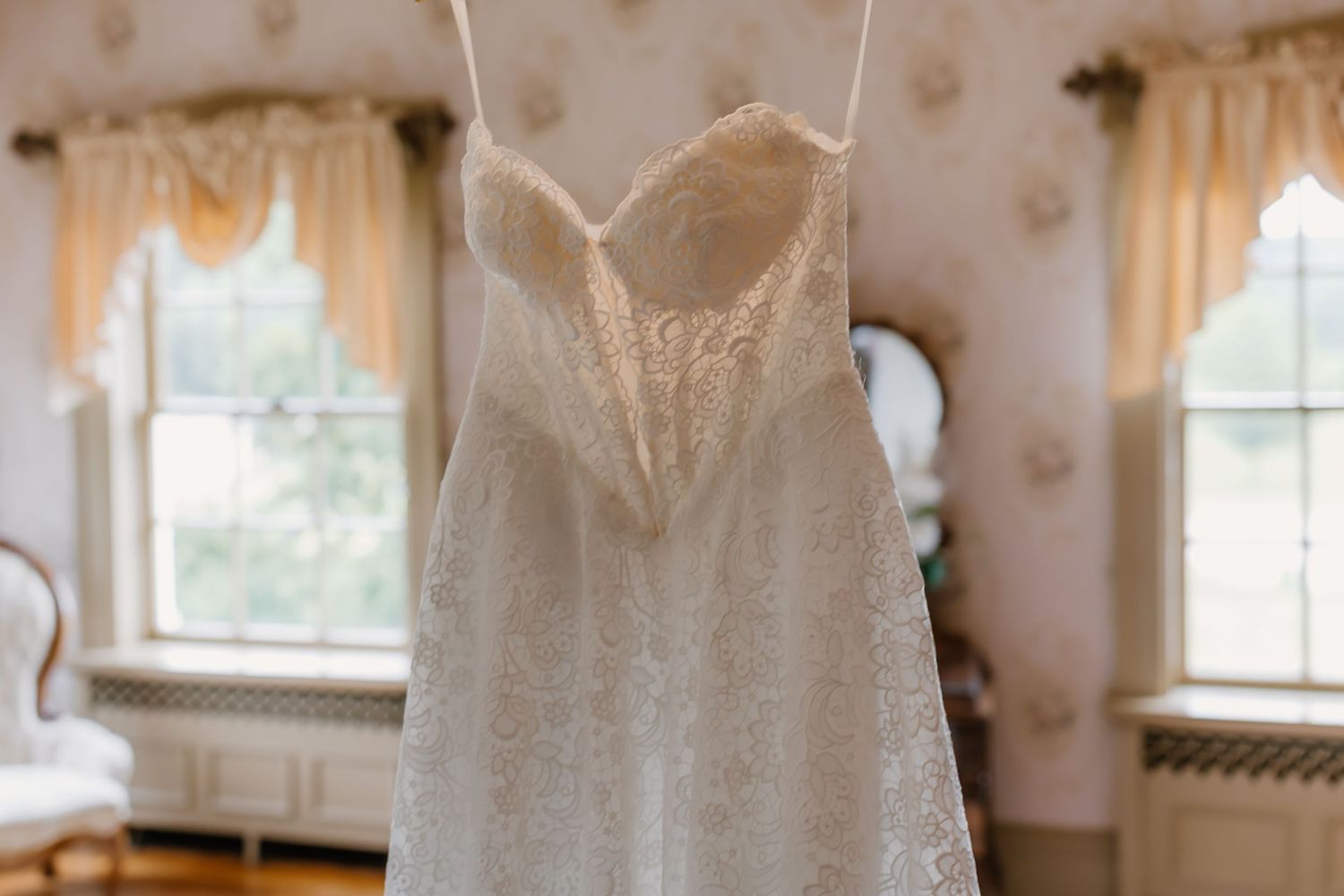 Elegant lace wedding dress hanging in a vintage room with floral wallpaper and soft natural light.