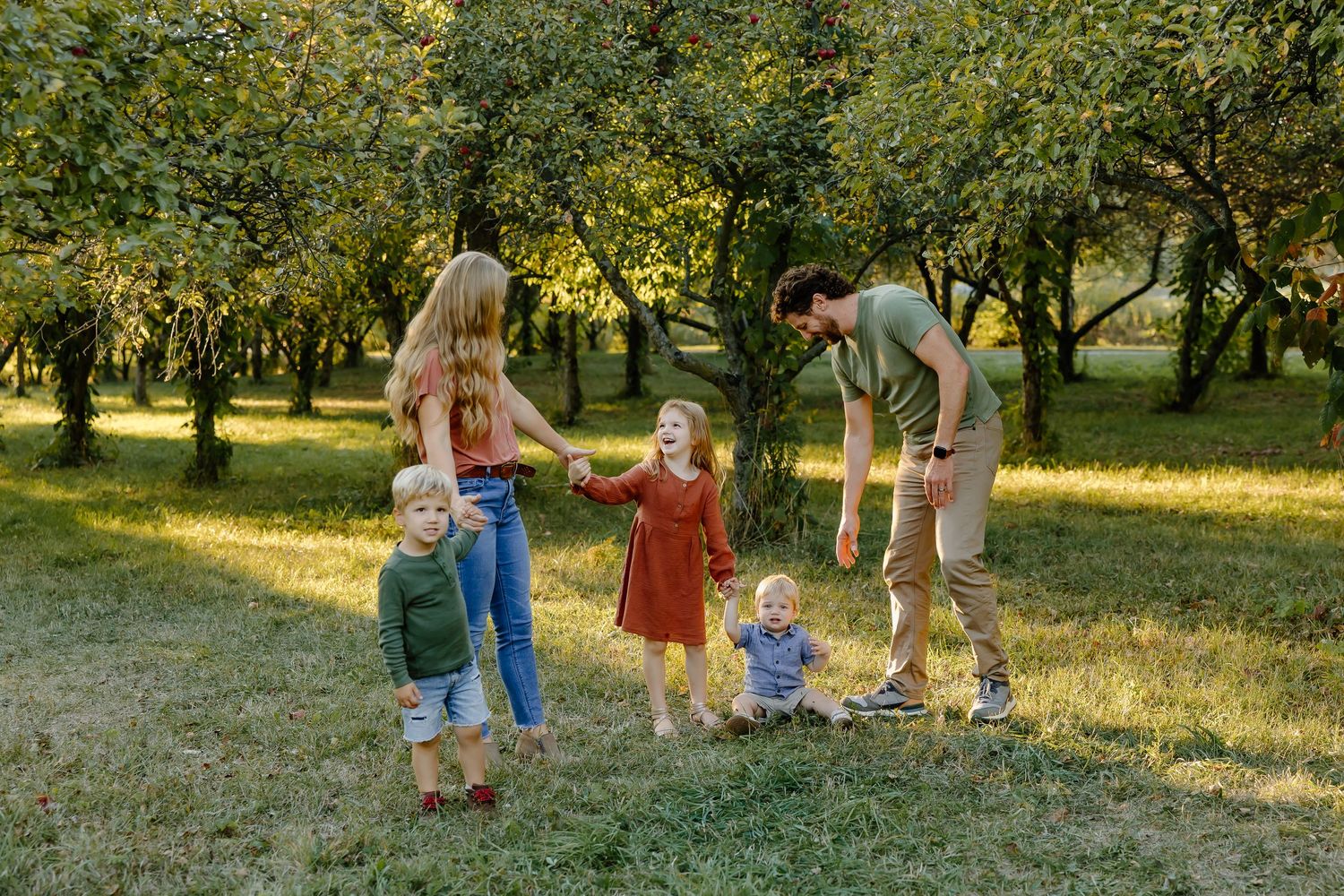 A family enjoying a playful moment together in a grassy orchard during sunset.