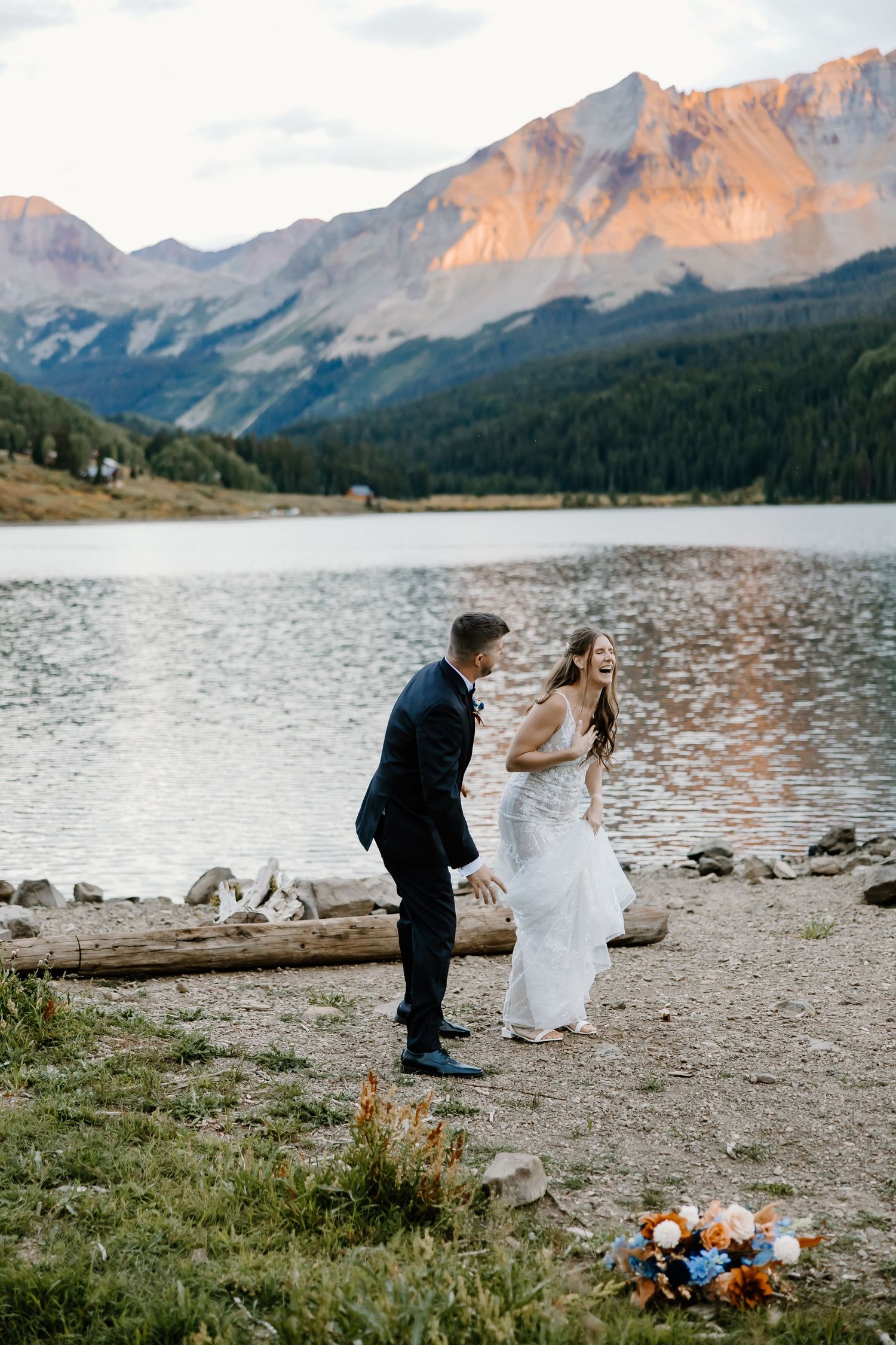 Couple celebrating by a mountain lake with wildflowers and driftwood at sunset.