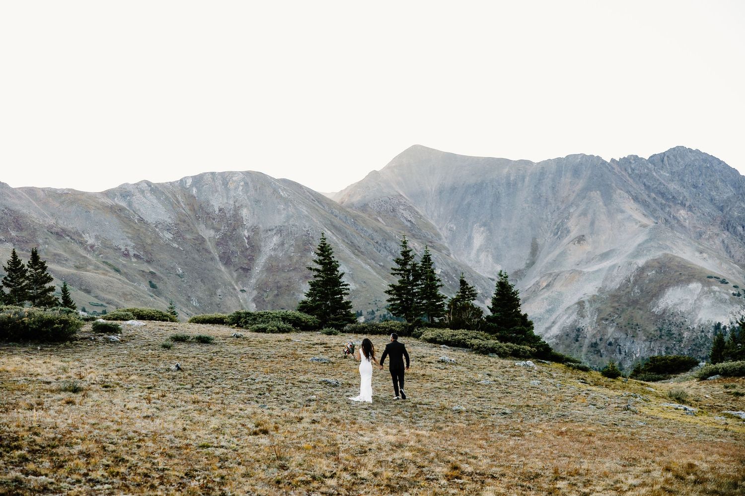 Distant view of wedding couple walking on mountain trail with dramatic alpine landscape.