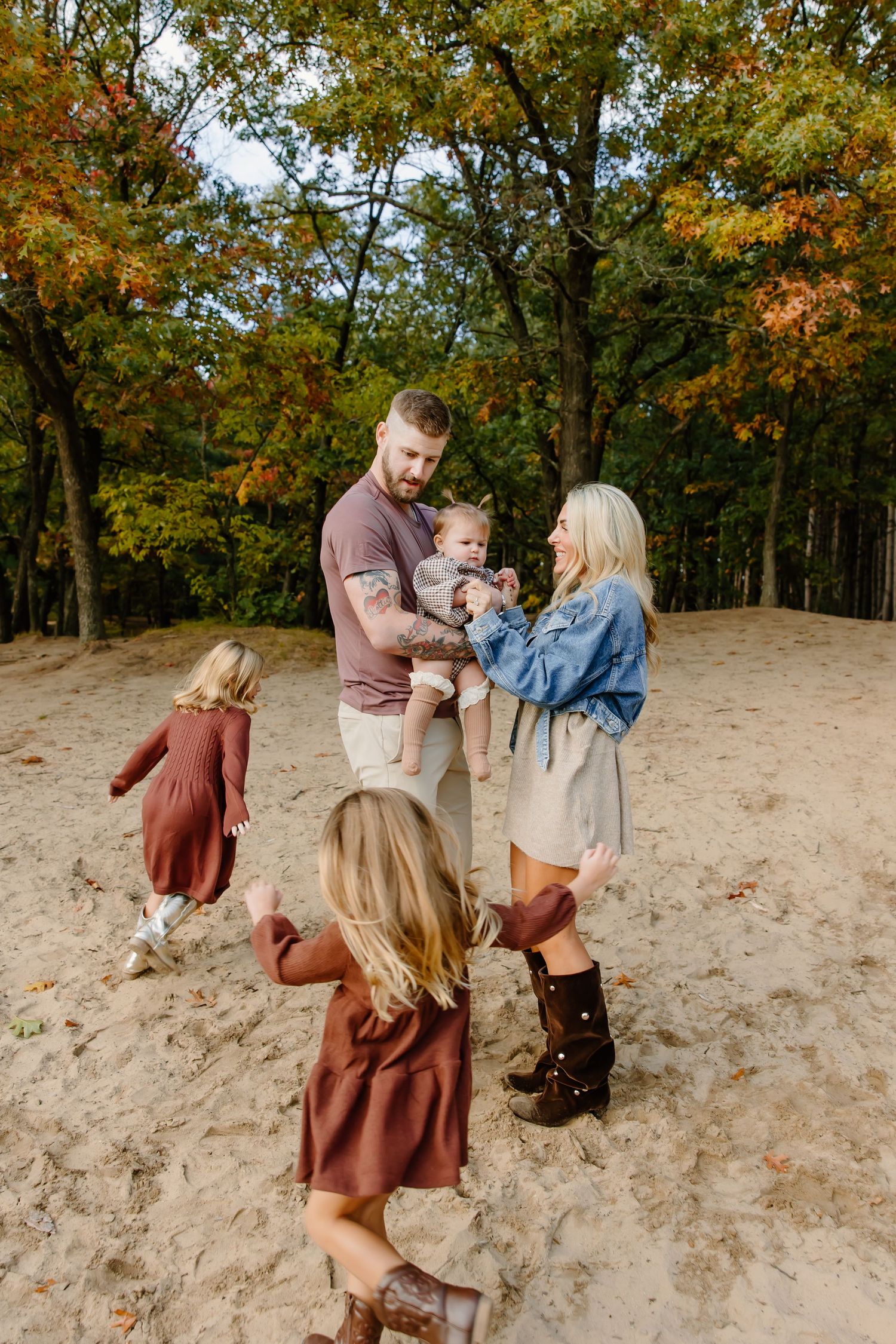 Family plays together on sandy hill surrounded by fall foliage in coordinated earth-tone outfits.