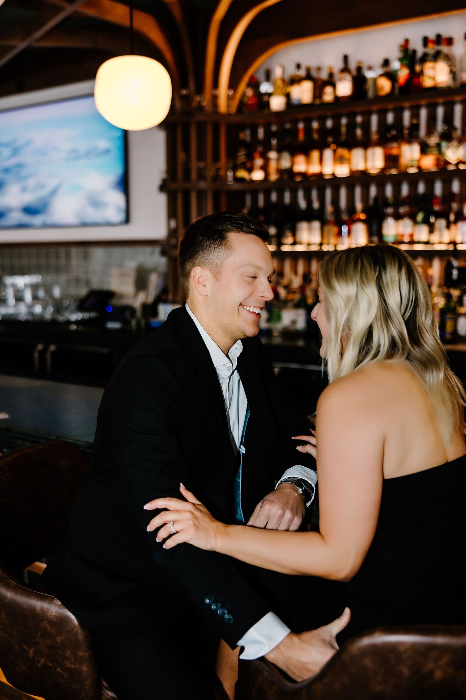 Couple sharing affectionate moments at a bar with bottles displayed in background.