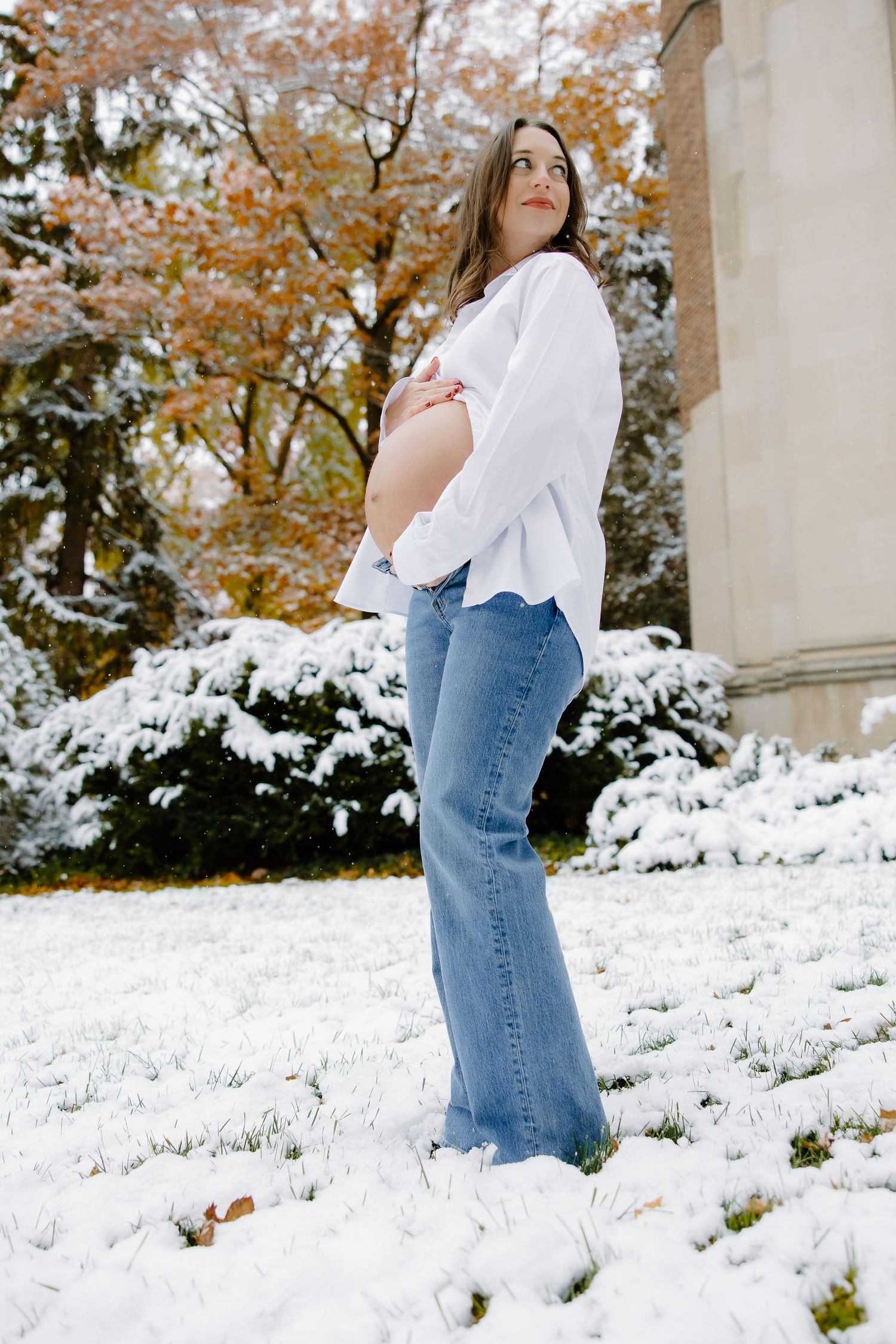 Maternity photo of woman in white shirt and jeans posing in snow with Gothic architecture.