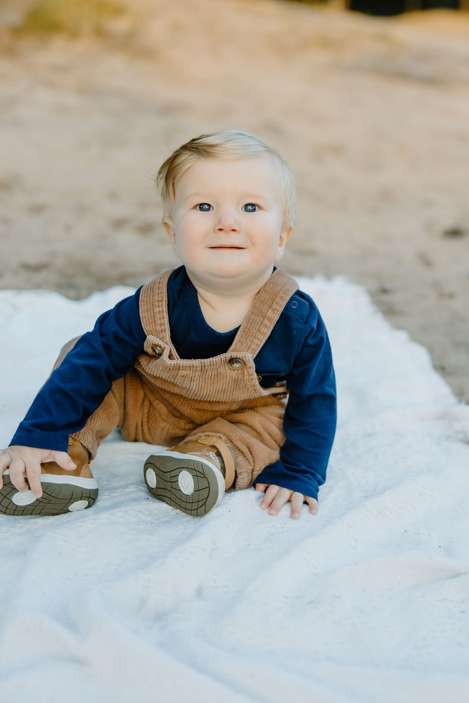 A baby wearing brown overalls and navy shirt crawls on a white blanket.