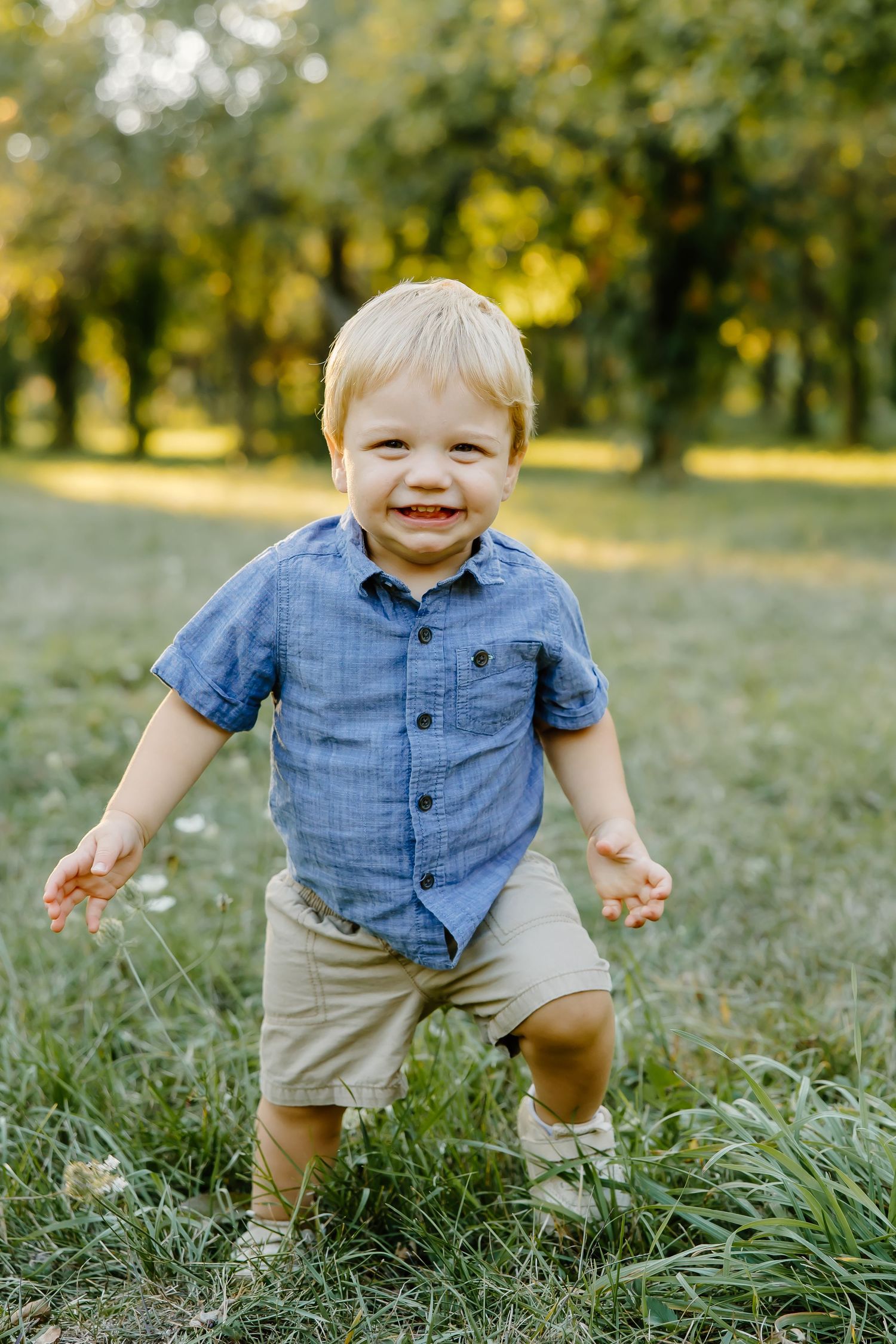 Toddler in blue button-down shirt and khaki shorts plays joyfully in a sunlit grassy field.