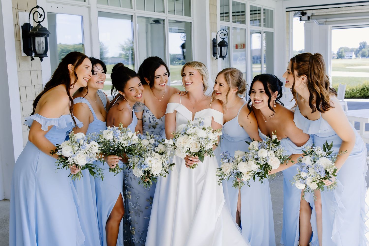 Wedding party members in light blue dresses pose with floral bouquets on a white porch.