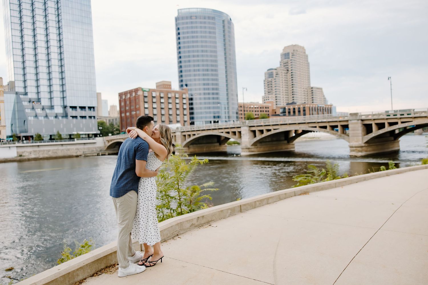 Couple sharing romantic moment by riverside with city skyline and bridge in background.