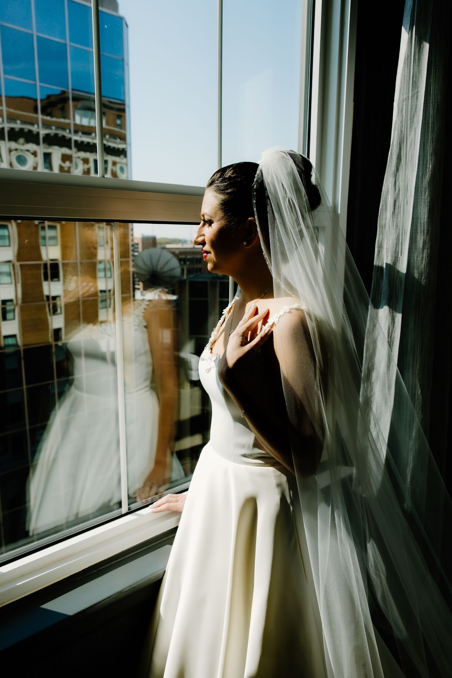 Bride in white dress and veil gazes through sunlit window.