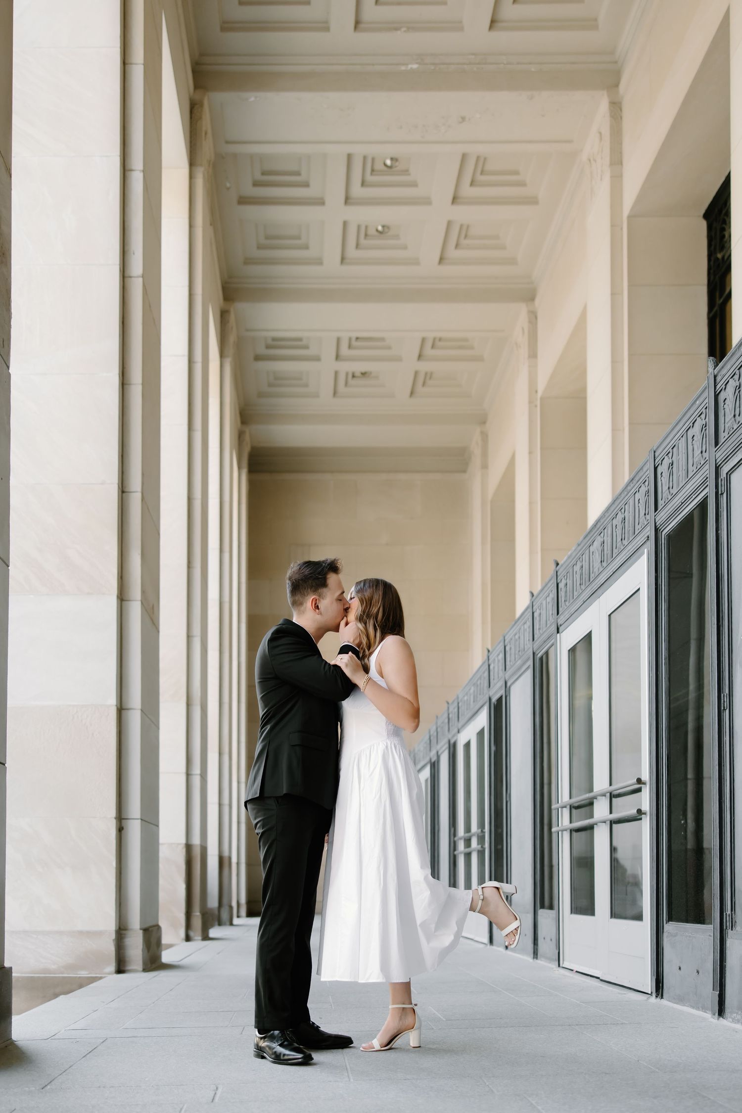 Couple sharing a romantic kiss in a grand architectural hallway, man in black suit and woman in white midi dress.