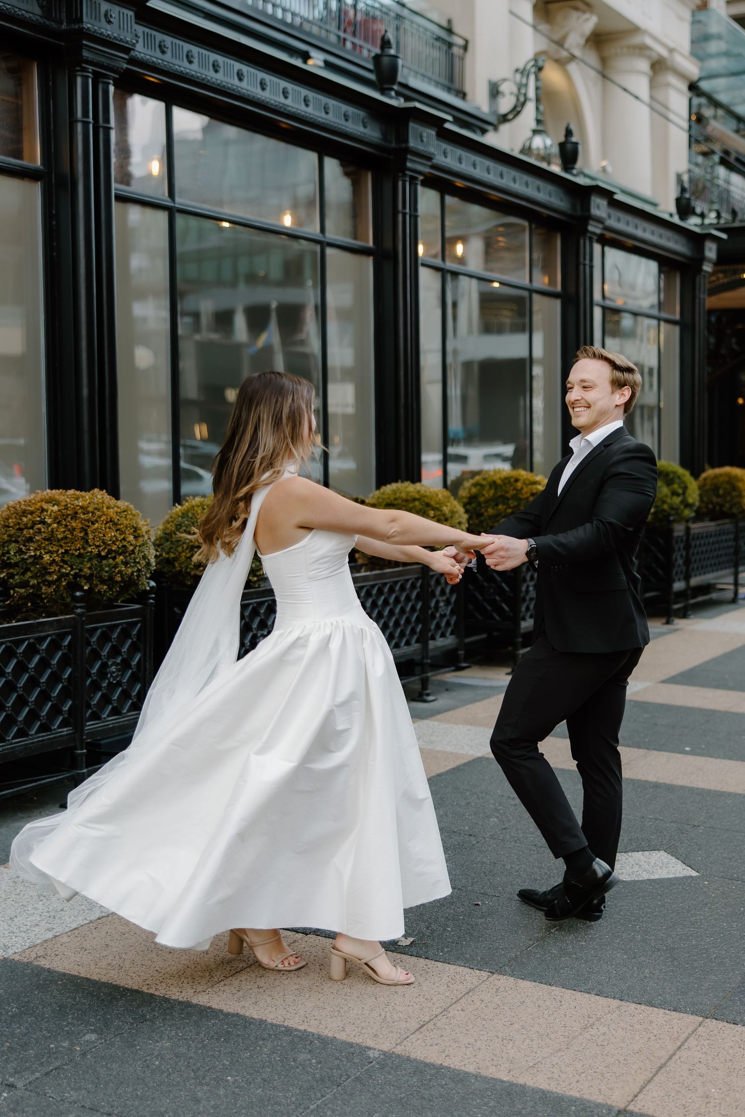 Bride in white dress and groom in black tuxedo dancing joyfully on a city sidewalk outside an elegant restaurant.