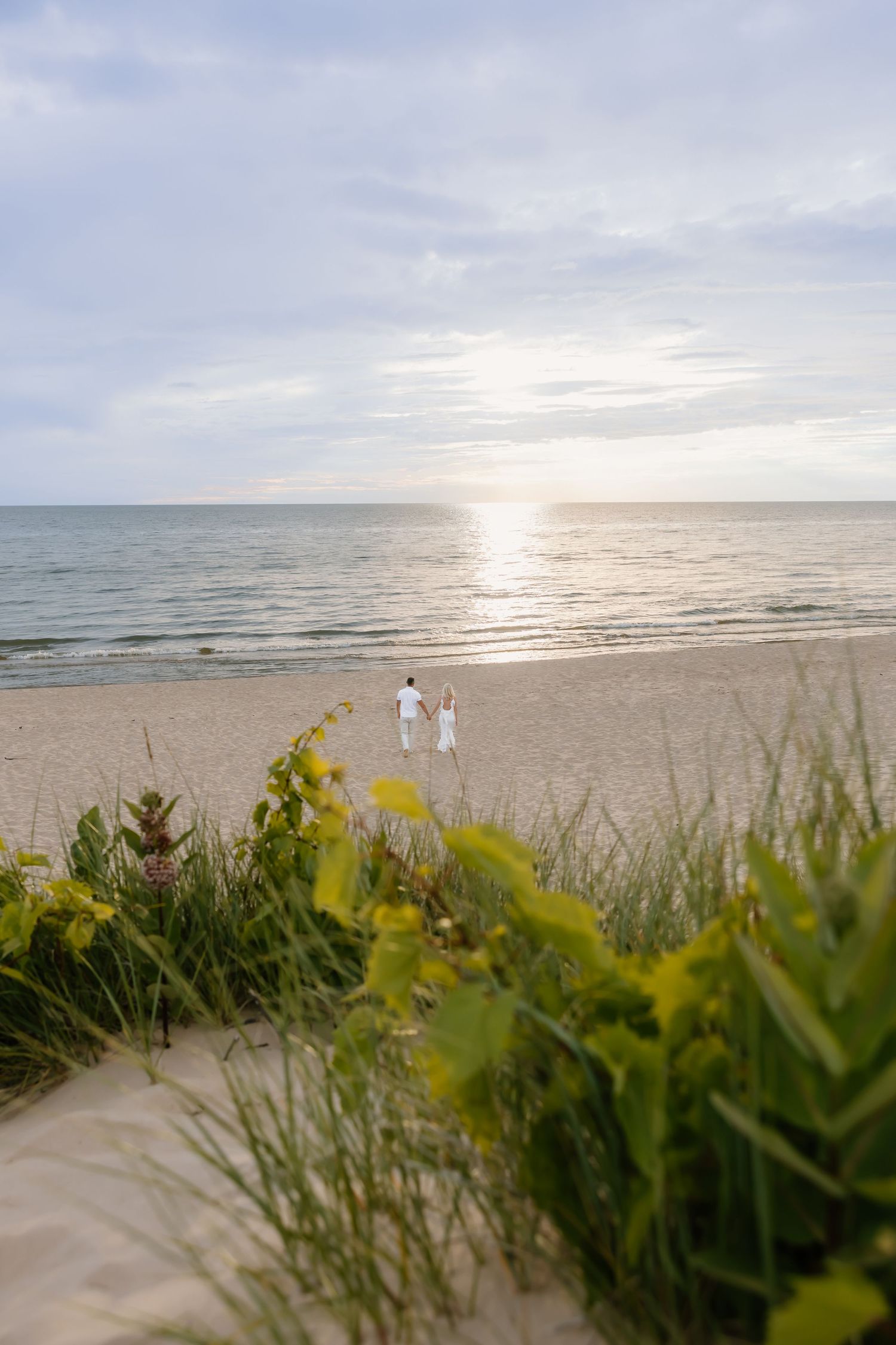 Couple walking hand in hand on a sandy beach at sunset, framed by green dune grass and wildflowers.