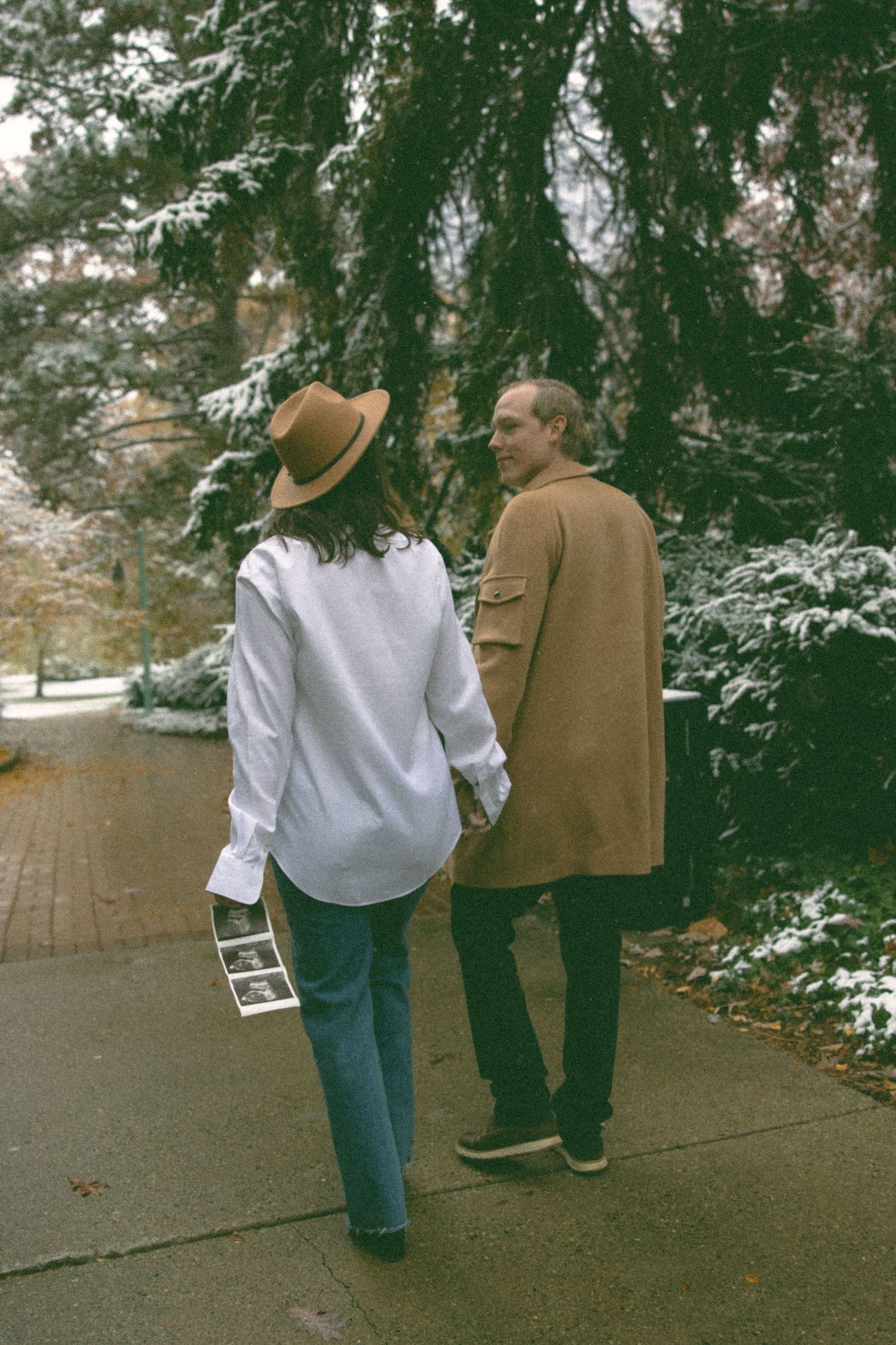 Couple walking together on a snowy path beneath evergreen trees.