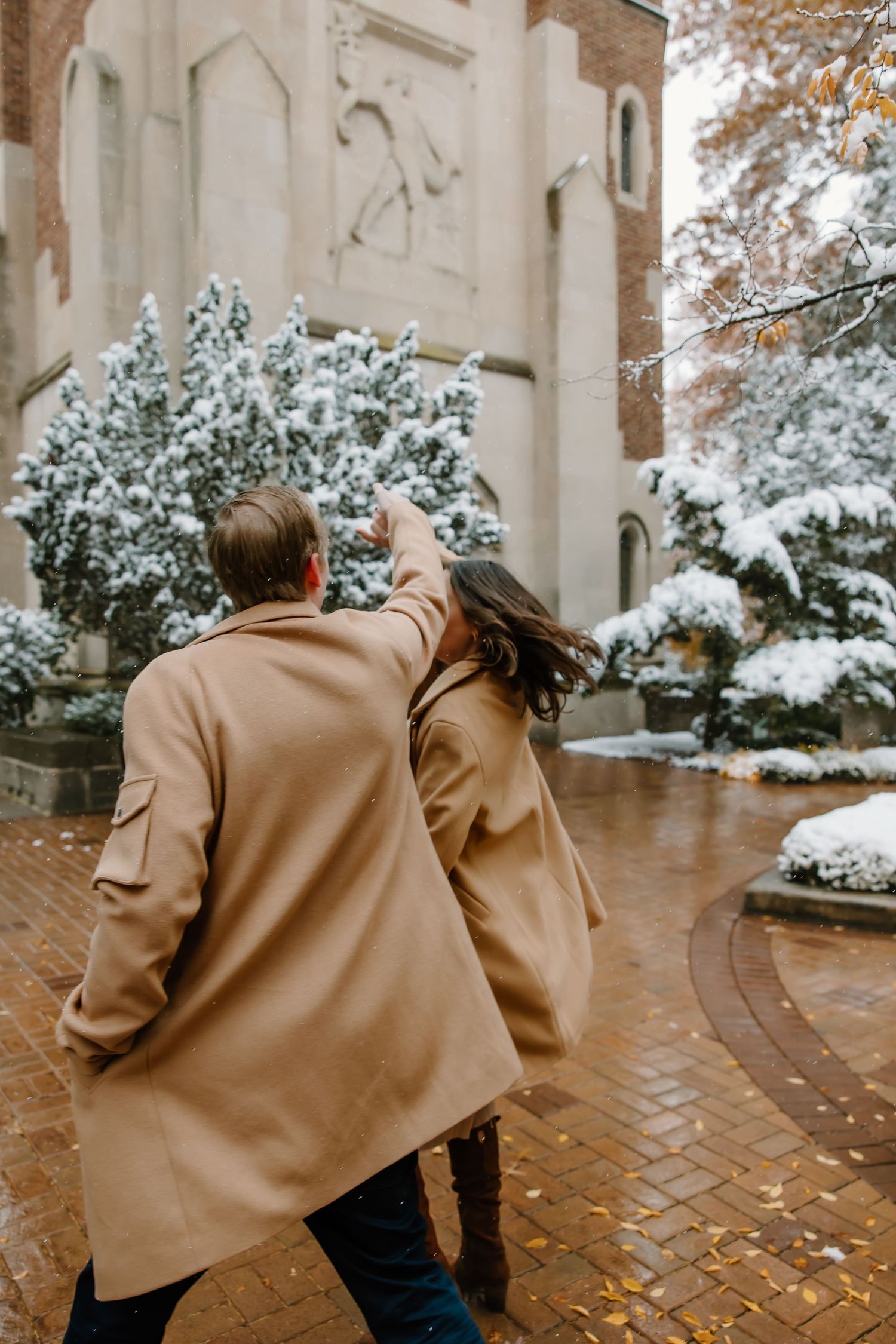 Couple in camel coats walking together on brick pathway near snow-covered gothic building.