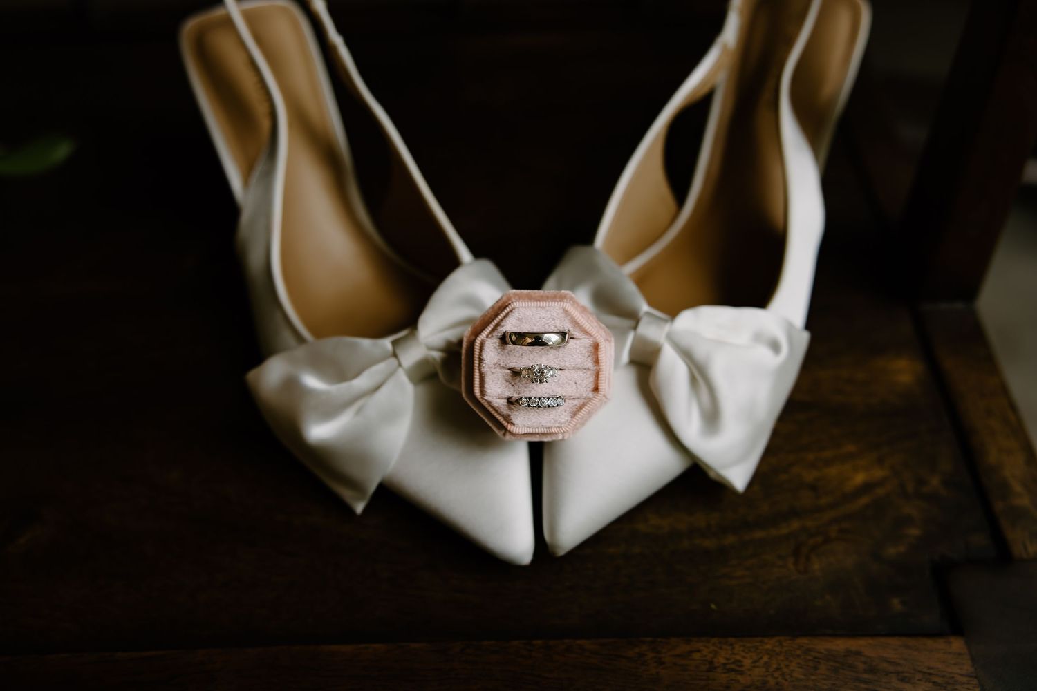 White bridal shoes and wedding rings arranged on dark wooden surface.