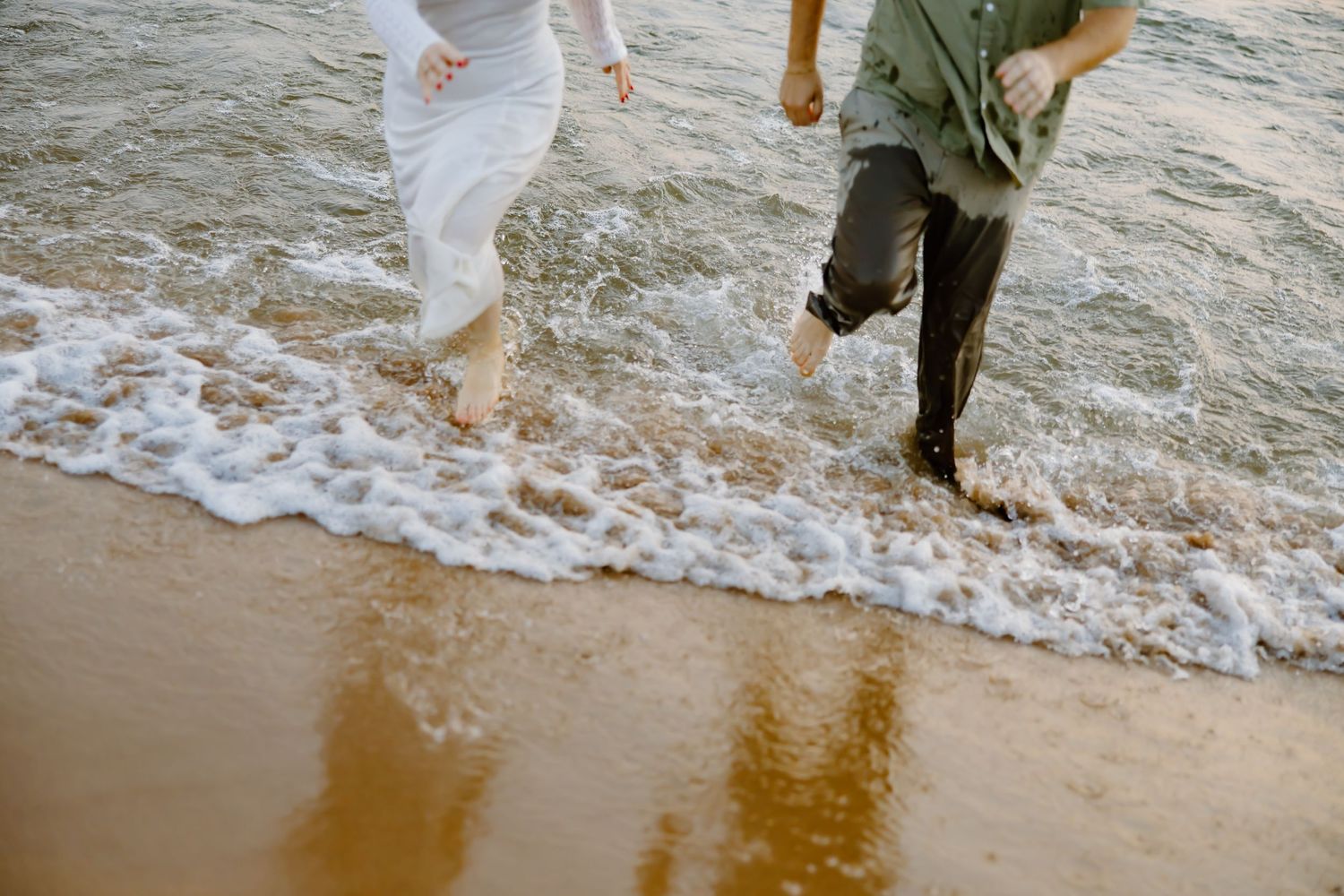 Couple's feet splashing in ocean waves on sandy beach at sunset.
